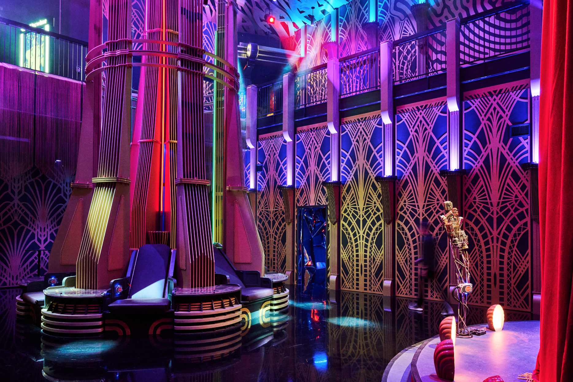 A colorful stage or club interior with vibrant neon lighting, geometric patterned walls, a large ornate chair, and a small statue figure near the right side, with red curtains on the far right.