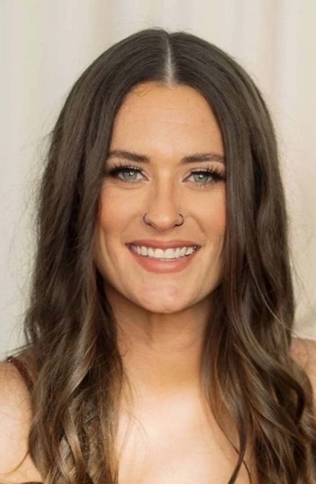 Close-up portrait of a smiling woman with long wavy brown hair, gray eyes, and a nose ring, against a neutral background.