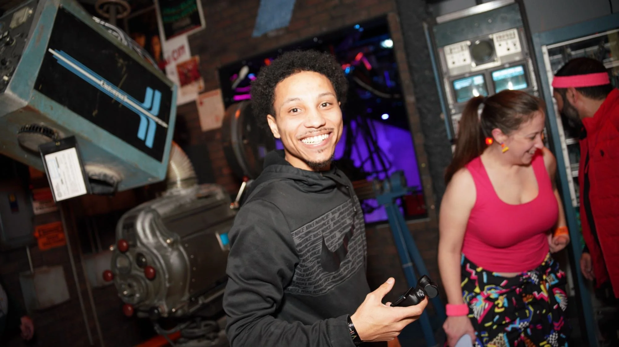 Smiling man in the themed projection booth of Atlas9 with two people in colorful outfits.