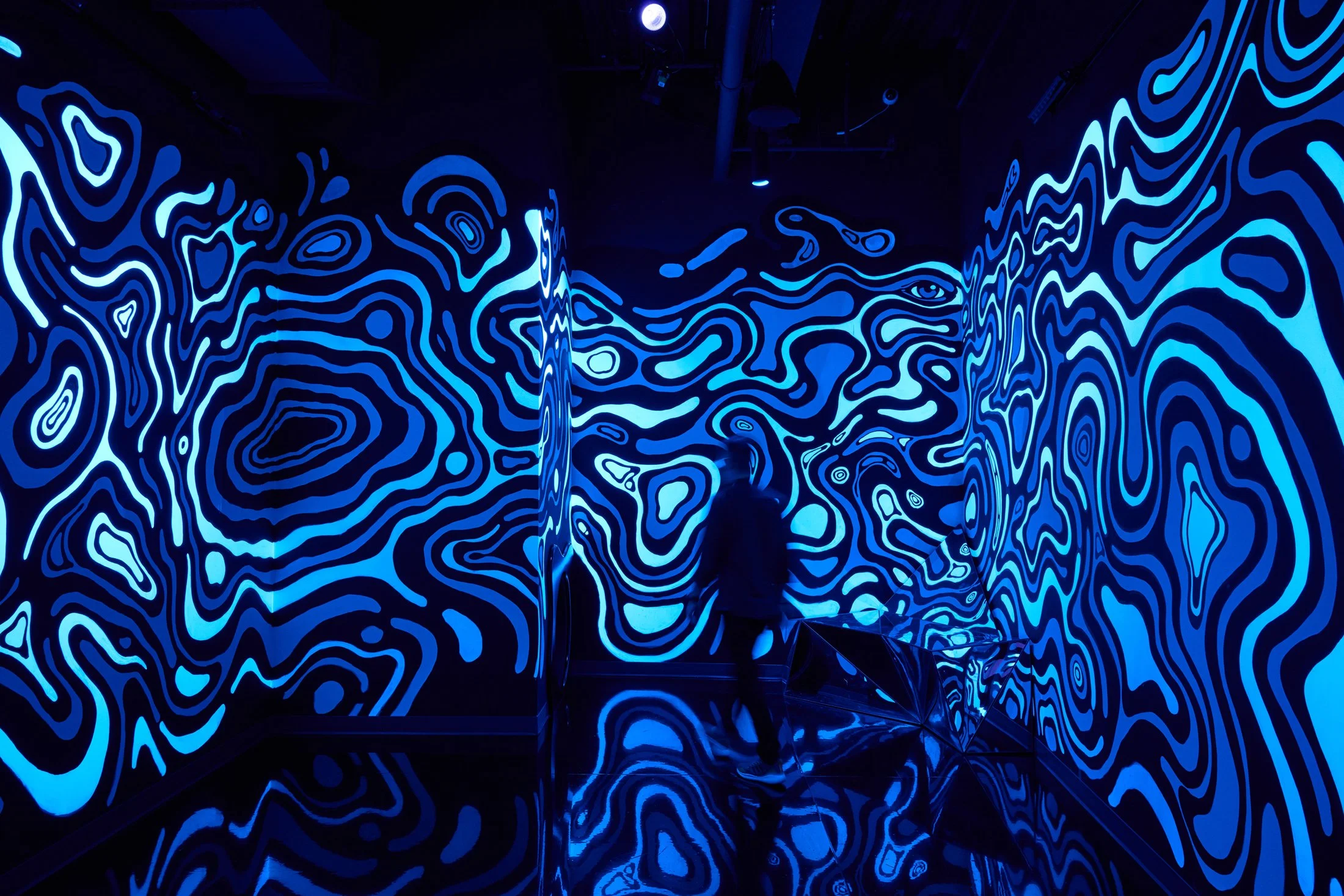 Person walking through a dark room with bright blue, neon, abstract swirling patterns on the walls and ceiling.