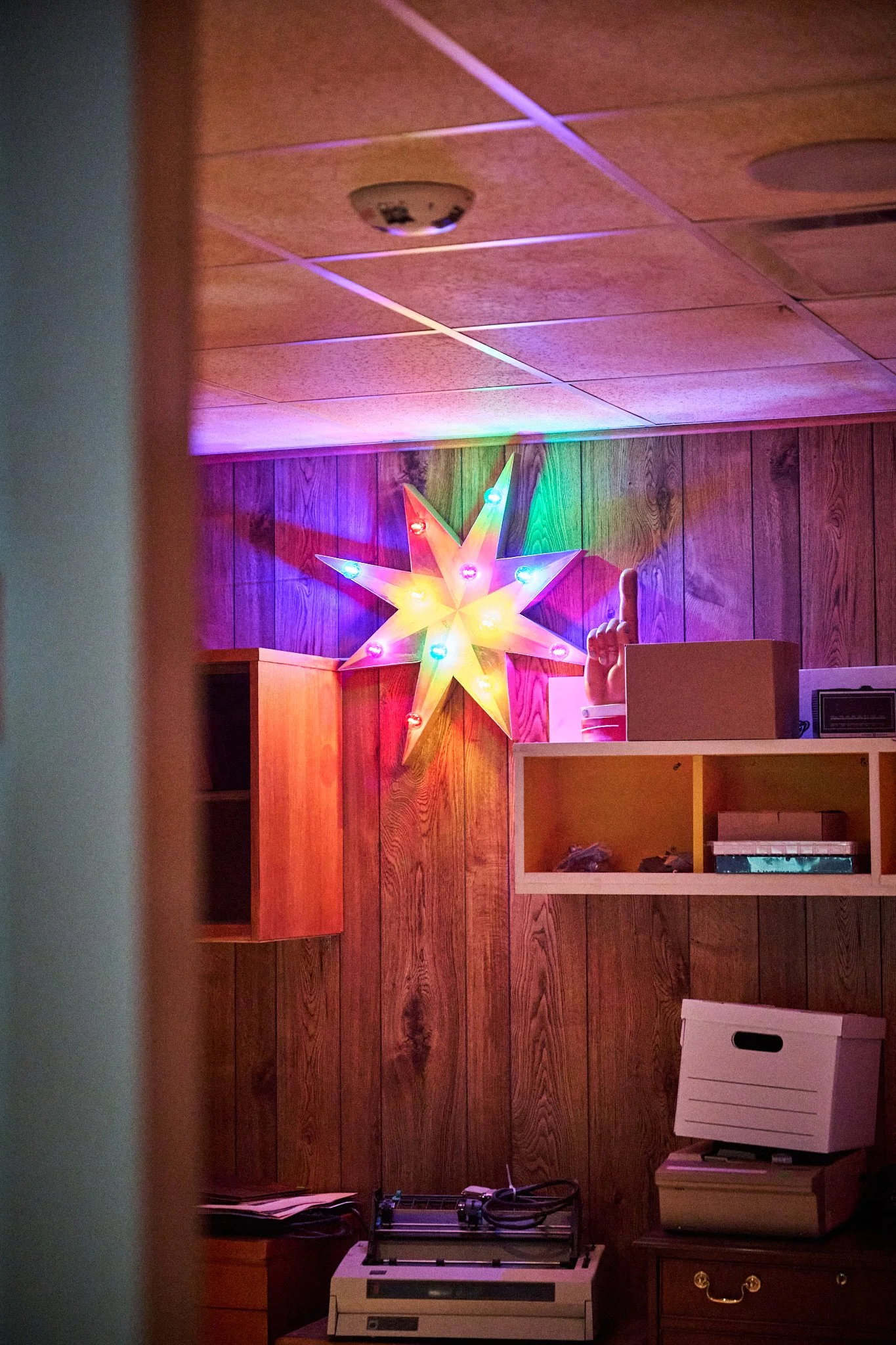 Wood-paneled room decorated with a brightly lit multi-colored star-shaped light on the wall, a mannequin hand with an index finger pointing upward, and various boxes and electronic equipment on furniture.