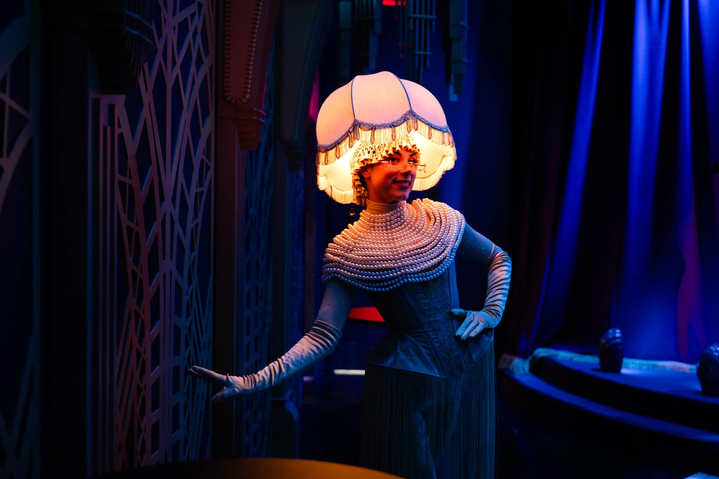 A performer dressed in a pink, fringed costume entertains an audience inside a colorful, illuminated jazz club theater with purple, pink, and blue lighting. She is in mid-air, holding a golden ball, with a lively pose.