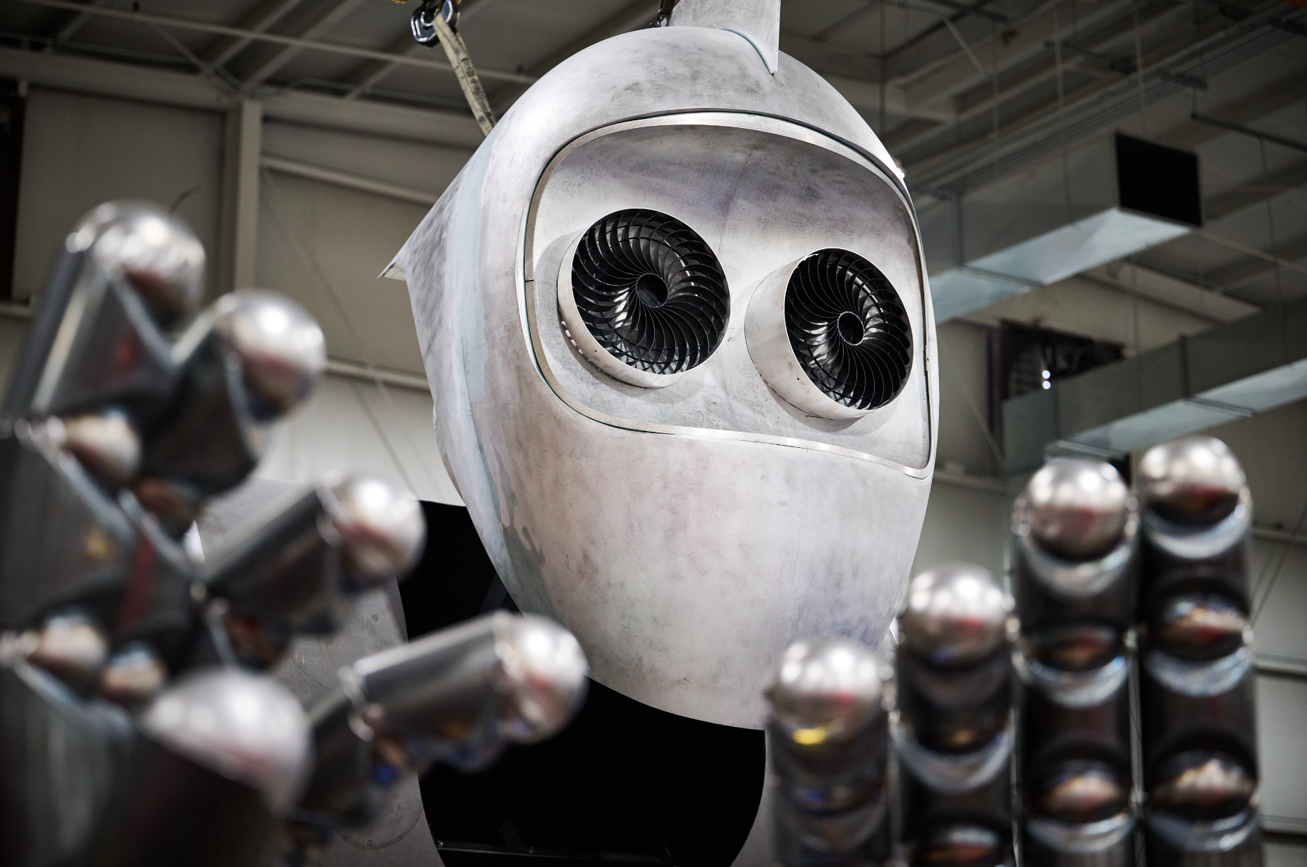 A robot for Atlas9 with a shiny, metallic face and round, black eye-like jet engines for eyes, surrounded by other metal robotic parts, inside an industrial or manufacturing setting at Dimensional Innovations.