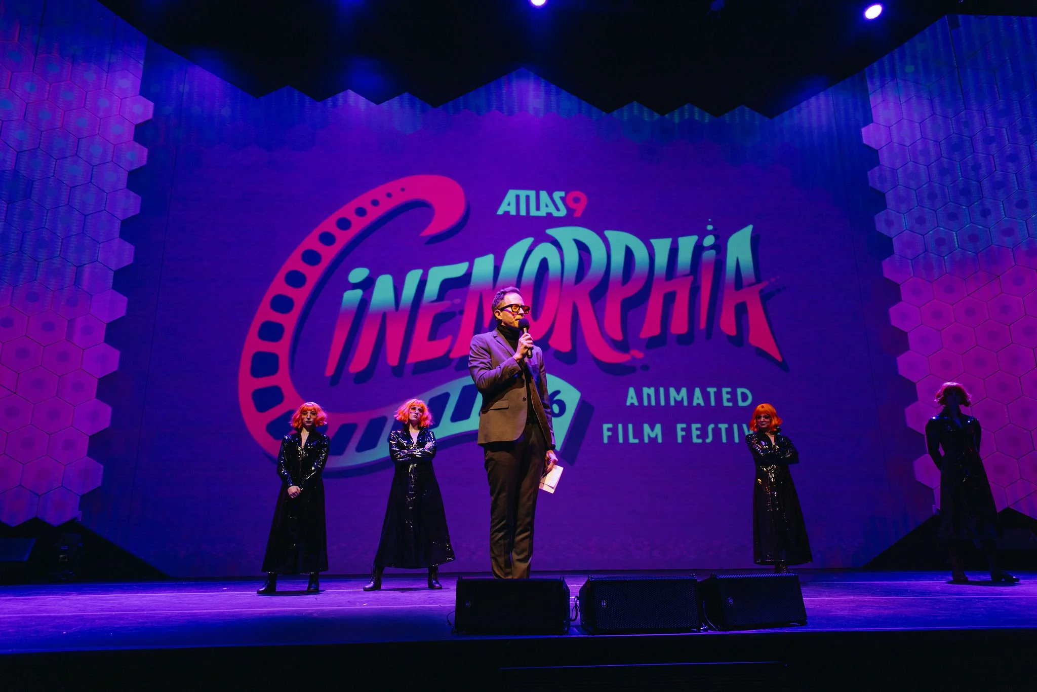 A man in a formal suit is speaking into a microphone on a stage at Atlas9, with four Quixotic women in shiny black dresses standing behind him. The backdrop displays the festival's colorful logo with the text CINEMORPHIA