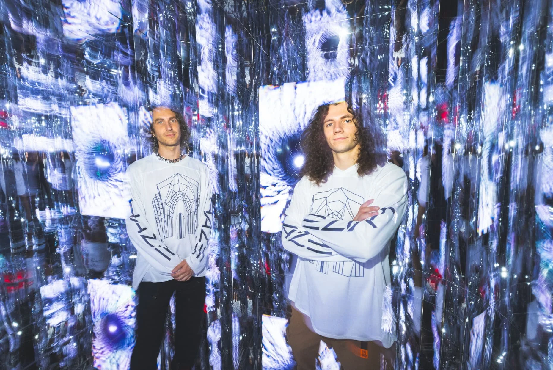 Two young men with curly hair standing amid reflective, illuminated silver walls, inside an immersive futuristic room at Atlas9 wearing white long-sleeve shirts with geometric designs.