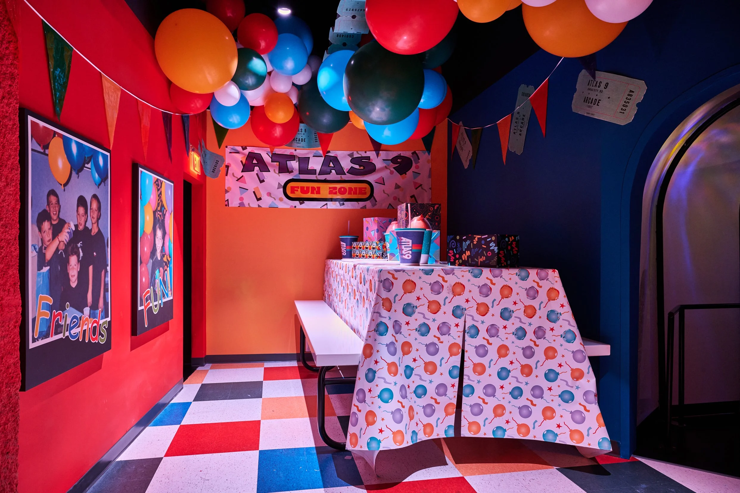 A colorful birthday party scene at an arcade or fun zone, decorated with multicolored balloons hanging from the ceiling, festive banners, and posters of children having fun. There is a table covered with a patterned tablecloth and a cake or gift boxe