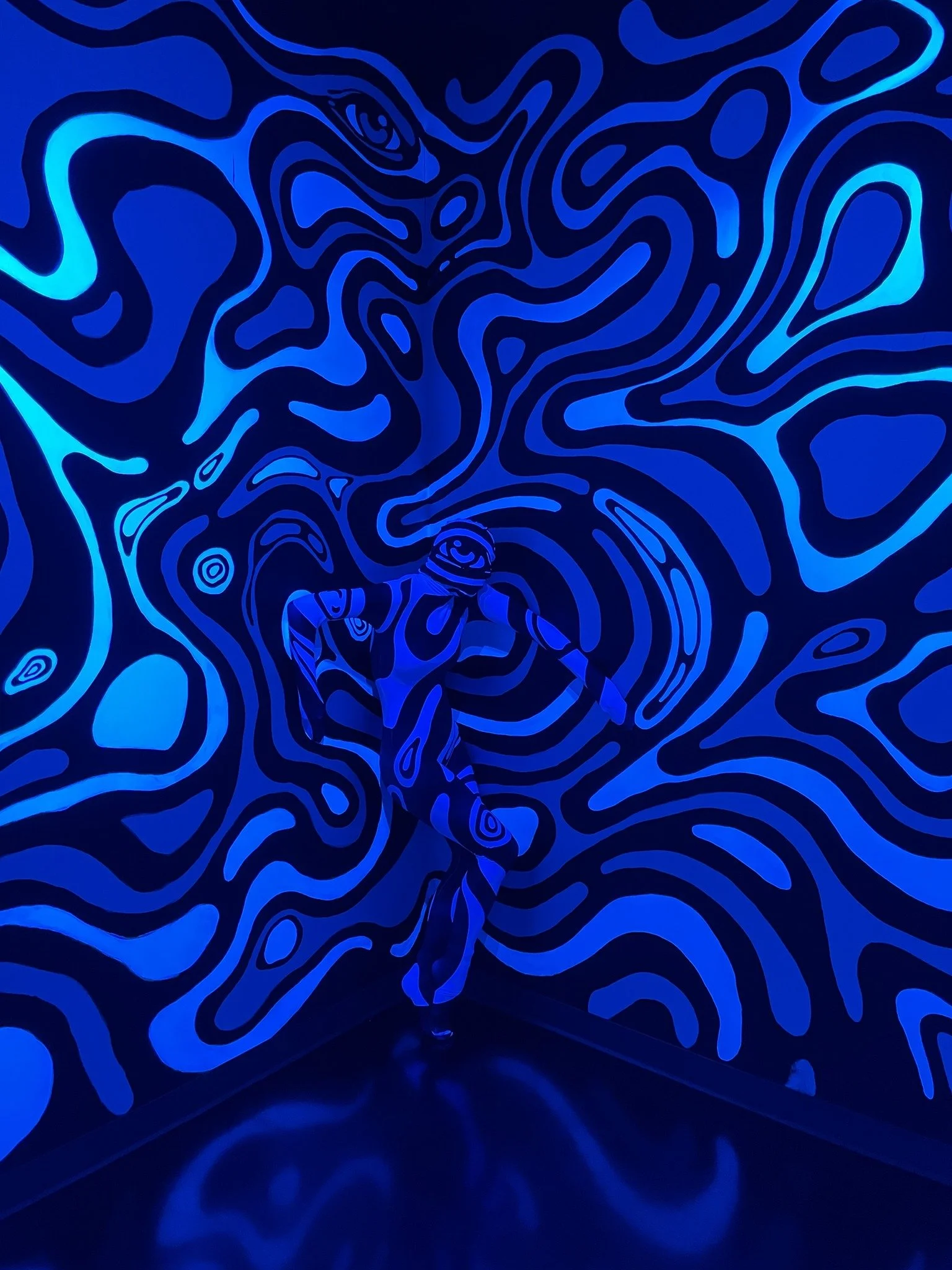 A Quixotic performer standing in a room with black and blue abstract, swirling, wavy line patterns on the walls and floor at Atlas9.