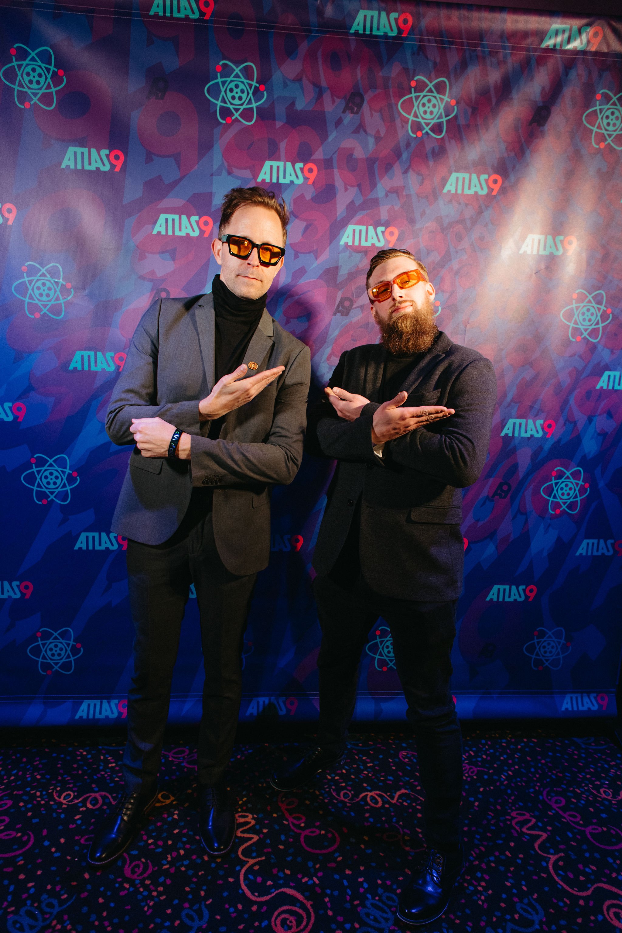 Two men with sunglasses posing in front of a vibrant backdrop with science-themed graphics and the word 'ATLAS9' repeated. They are dressed in suits and stand with crossed arms pointing at each other.