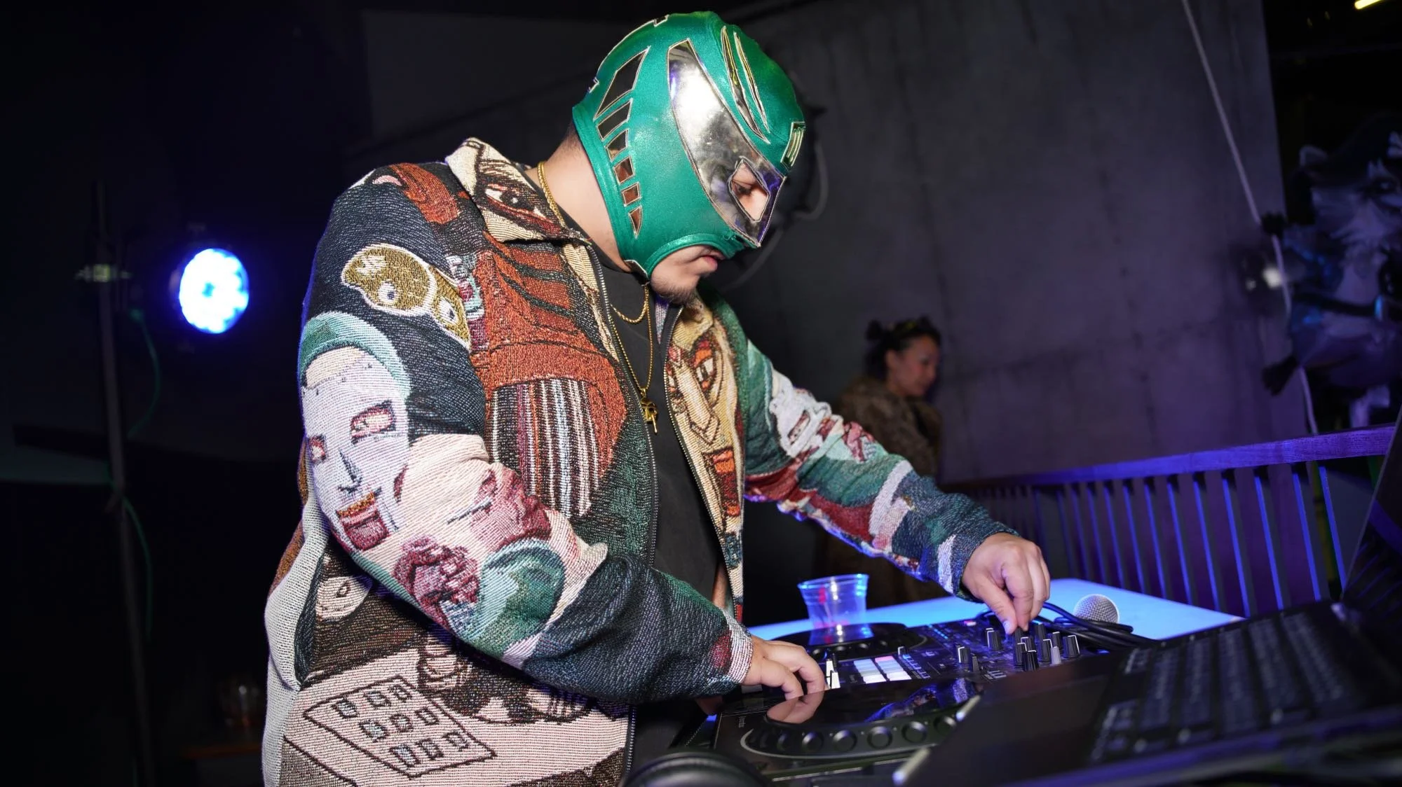 DJ wearing a green helmet and colorful jacket mixes music at a stage with purple and blue lighting.