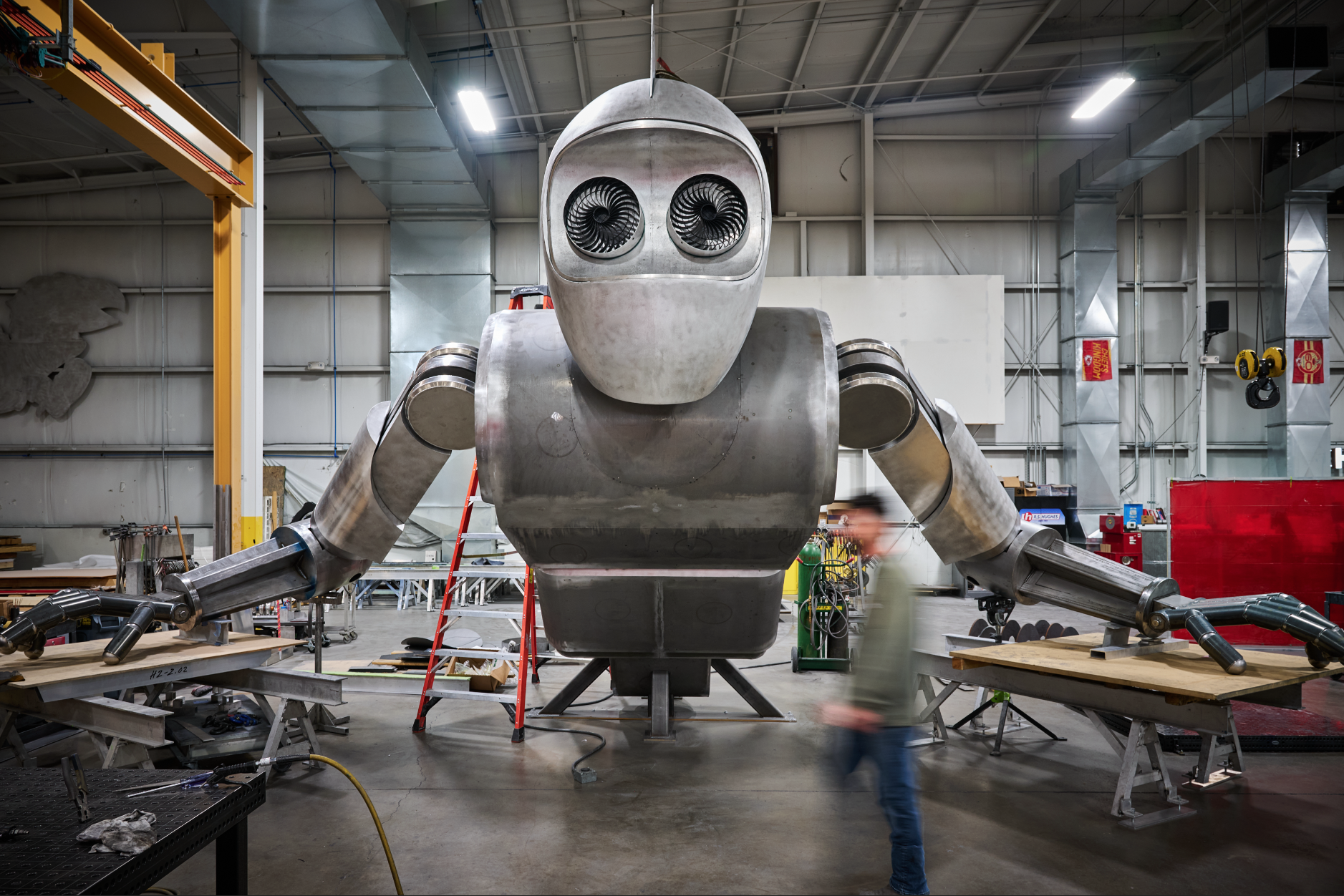 A large humanoid robot getting built for Atlas9 with a metallic body and rotary fans for eyes inside a spacious industrial workshop at Dimensional Innovations