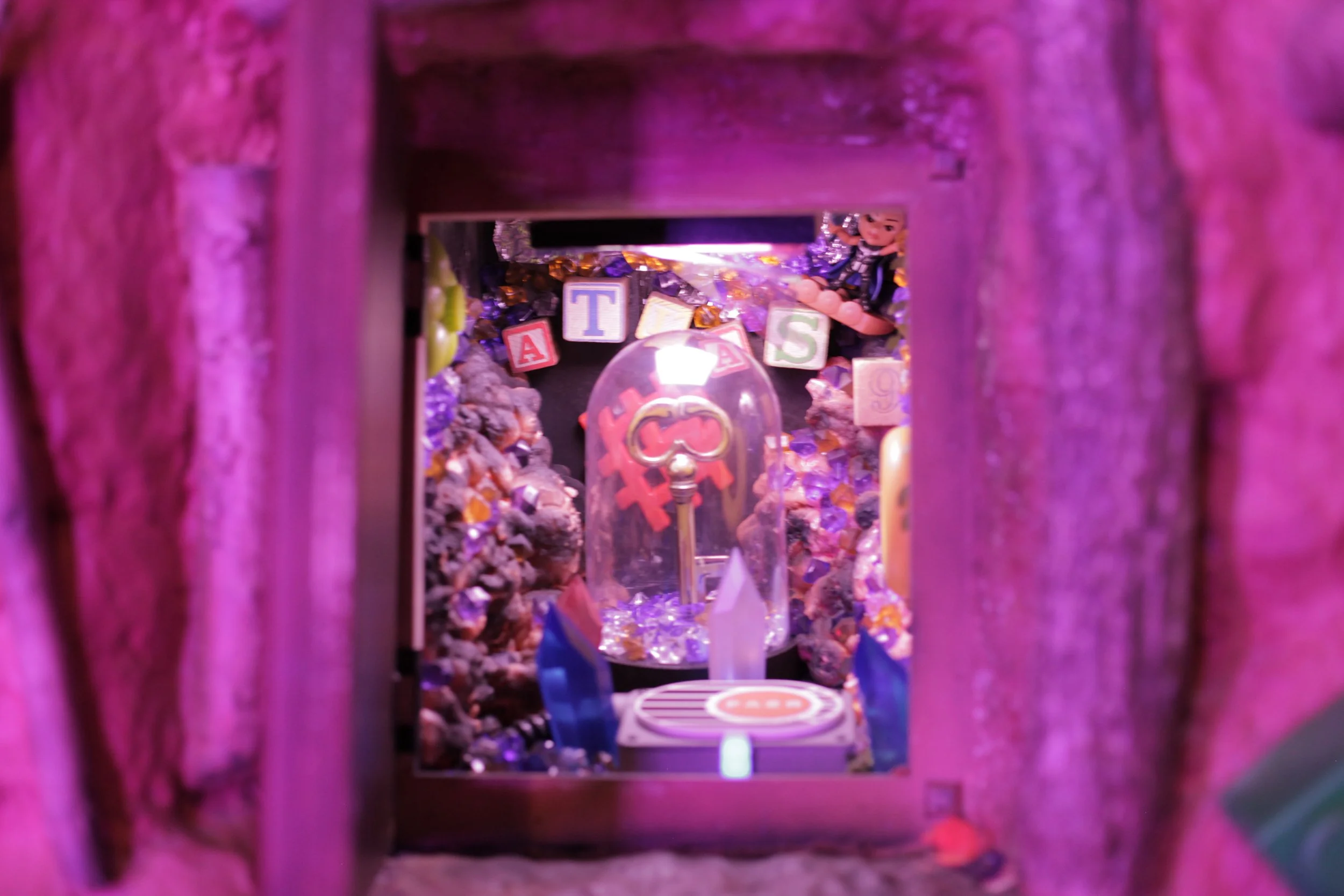 A colorful diorama featuring a small figure, a key under a glass dome, and alphabet blocks spelling 'TAS,' surrounded by rocks and decorations.