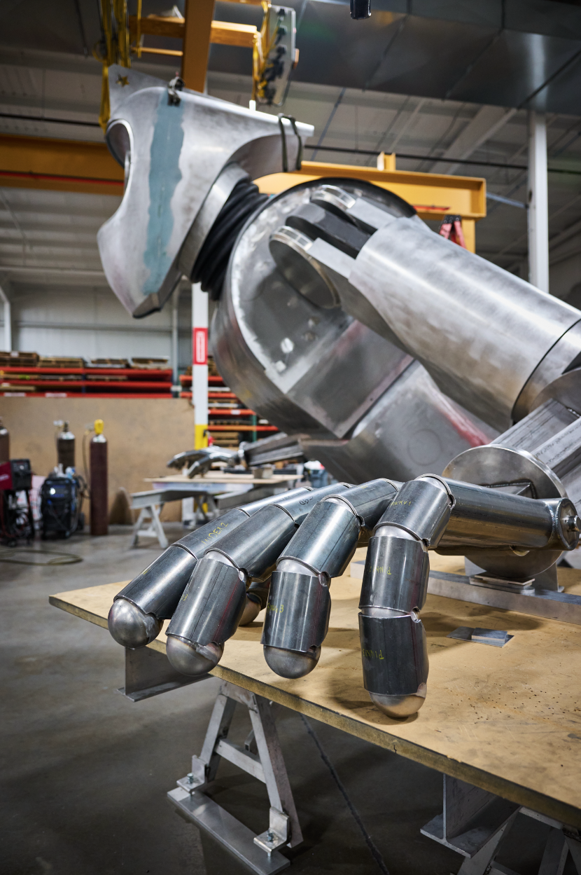 A robotic hand made of metal parts resting on a workbench in an industrial workshop at Dimensional Innovations, with a large metallic robotic arm in the background for Atlas9.