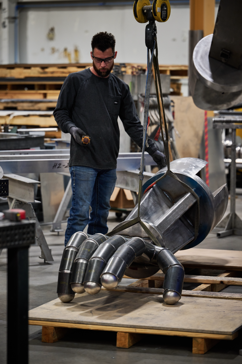 A man in safety glasses and black gloves is working with a crane to lift a large, shiny metal sculpture for Atlas9 resembling a robot with multiple legs, in a workshop at Dimensional Innovations surrounded by tools and wooden pallets.