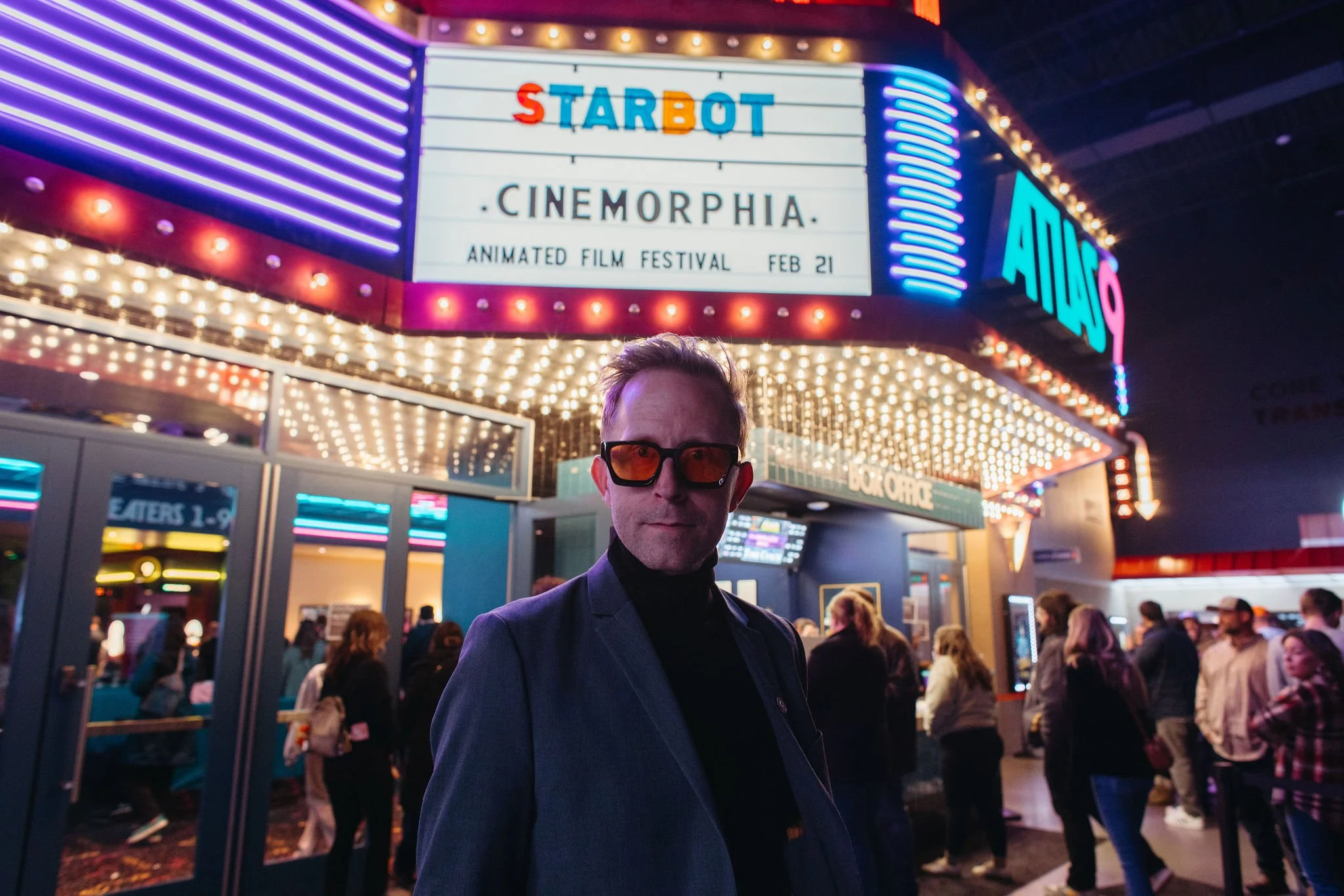 A man wearing sunglasses and a blazer stands in front of a crowded entrance to a film festival at night. The background features brightly lit signage for 'Cinemorphia' at the Animated Film Festival inside Atlas9