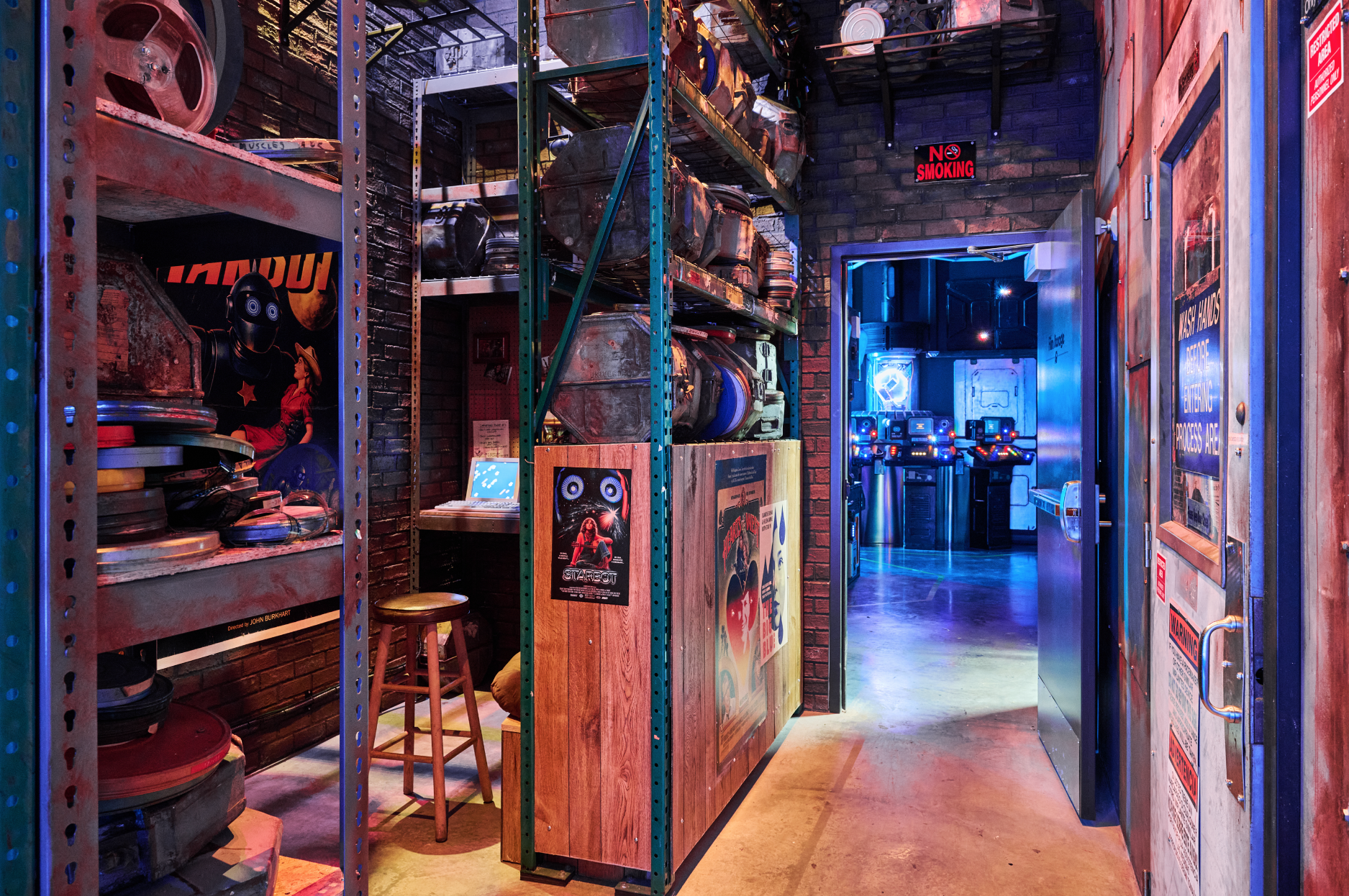 A hallway leading into a game arcade with shelves of vintage VHS tapes and posters on the walls. The arcade area is illuminated with neon and blue lighting, with arcade machines visible in the background.