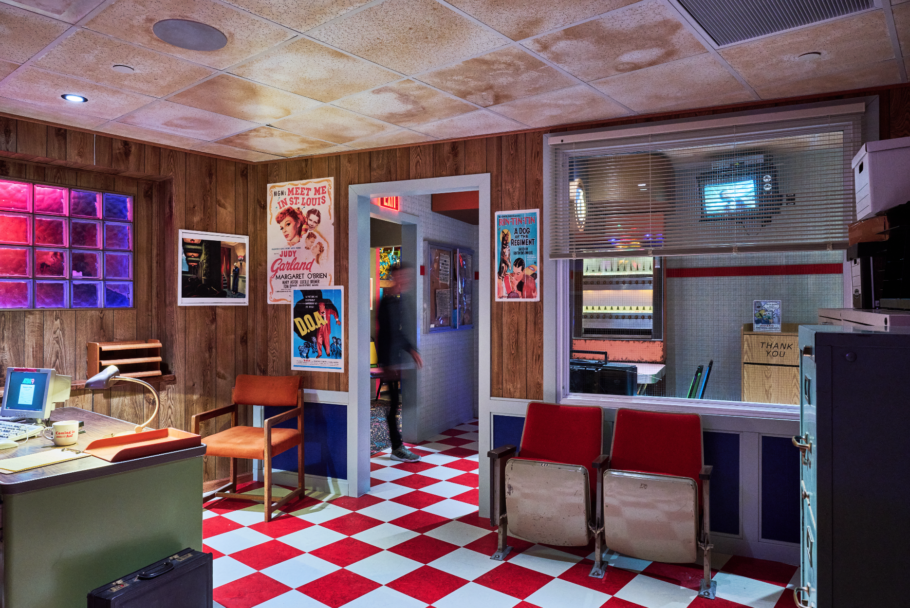 Interior of a vintage movie theater lobby with wood-paneled walls, red and white checkered flooring, posters on the wall, a desk with a lamp, and red seating.