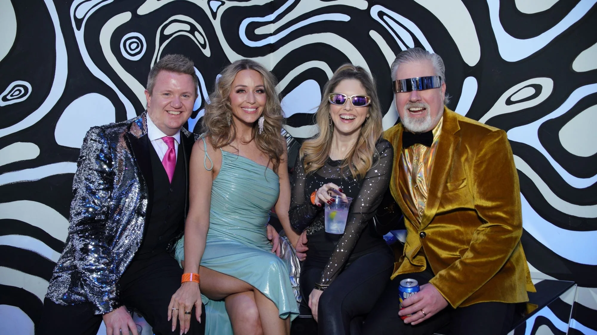 Four people in colorful, shiny, and patterned outfits at a party, sitting against a black-and-white abstract wall, smiling and holding drinks.