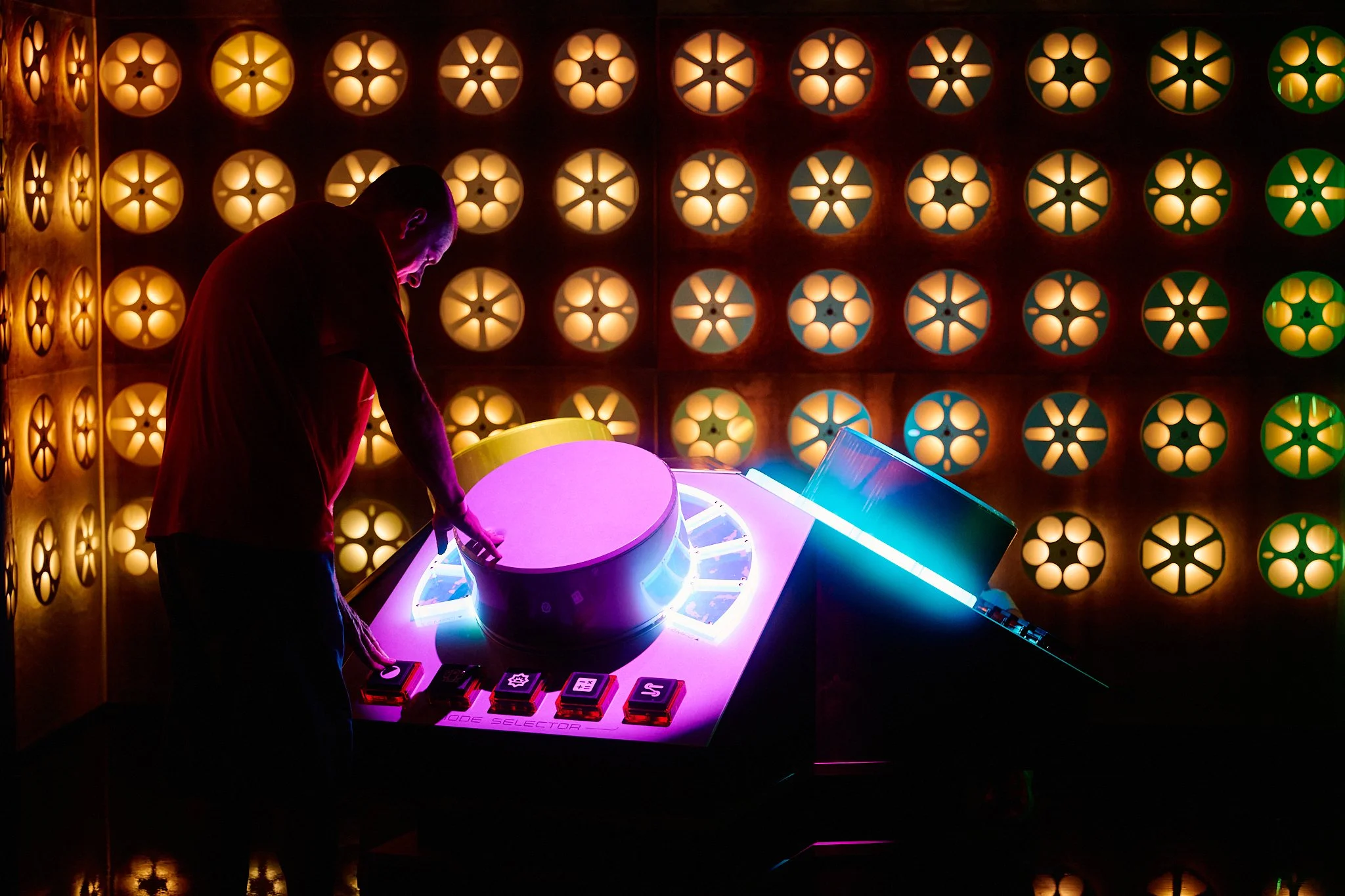 A person interacts with a futuristic, illuminated control panel in a dark room with a colorful, patterned wall background inside of Atlas9.