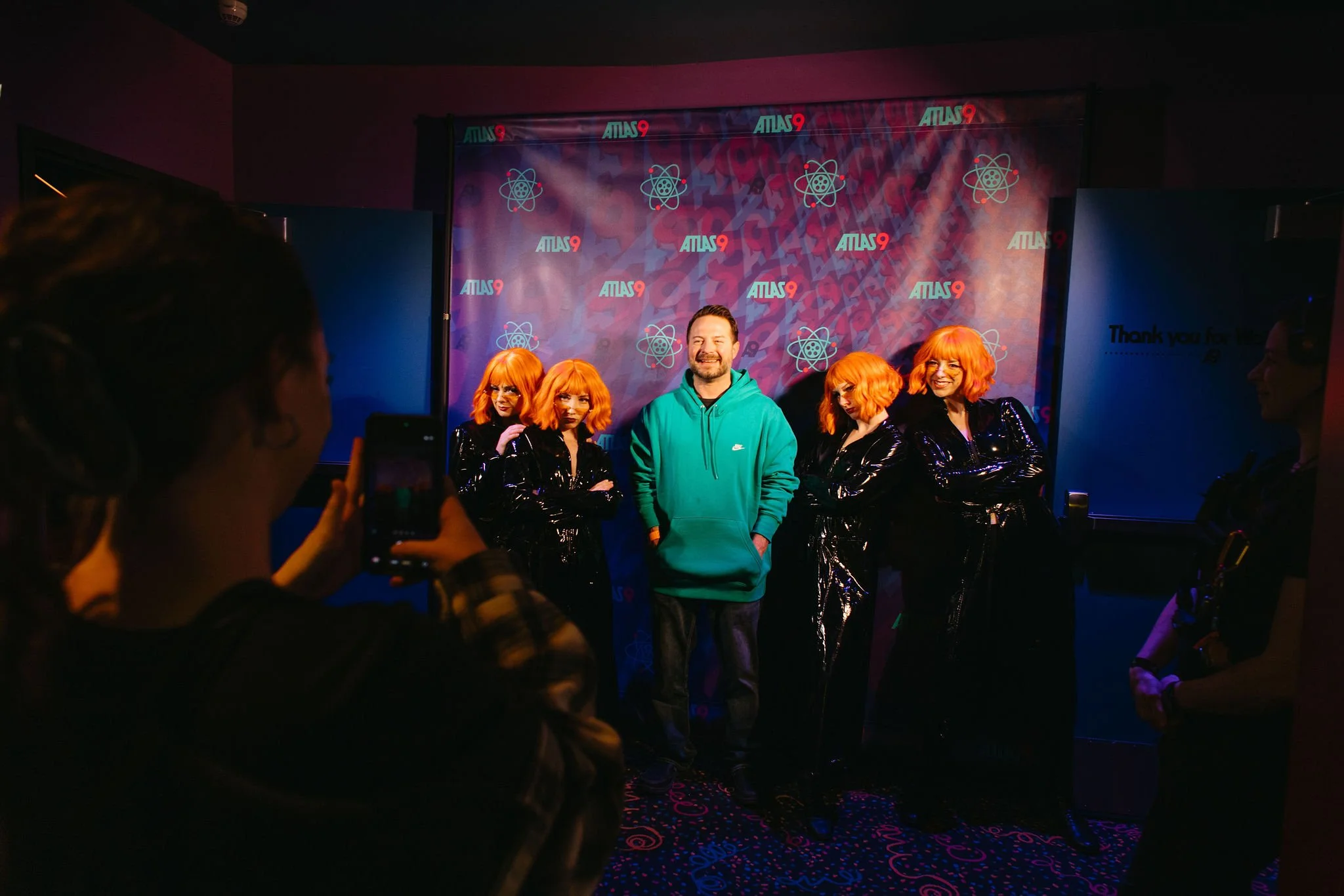 Group photo of five women with orange hair wearing black shiny outfits and a man in a teal hoodie standing in front of a backdrop that says 'ATLAS 9' with atomic symbols, as someone takes a picture.