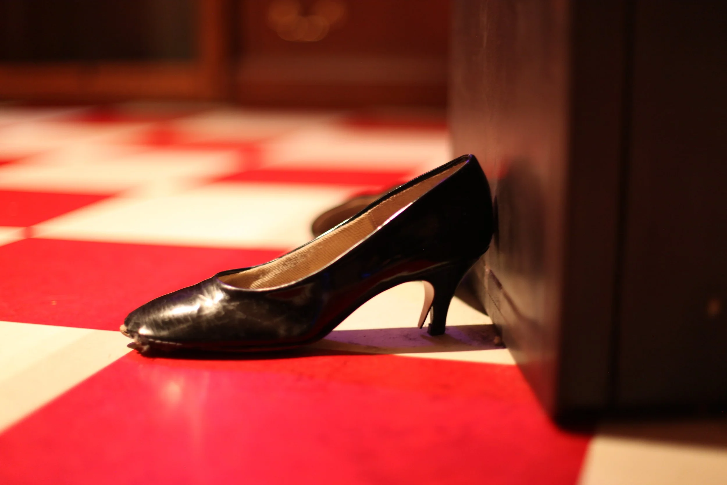 Black high-heeled shoe resting on a red and white checkered floor, with a wooden piece nearby and blurred background.