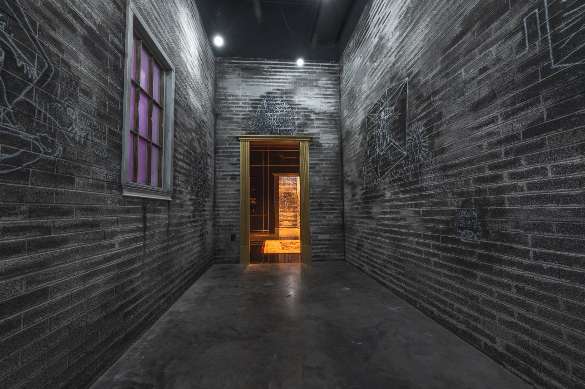 Dark indoor hallway at Atlas9 with brick walls decorated with chalk drawings, a window on the left with purple lighting, and a doorway at the end showing a warmly lit room with a rug inside.