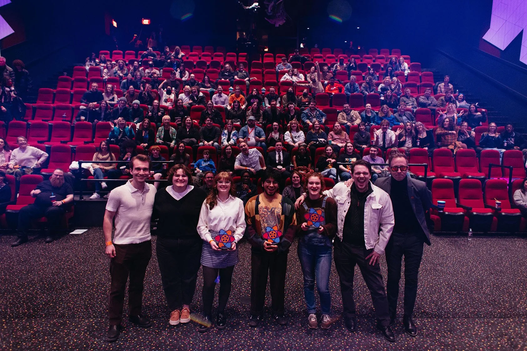 Group of seven people standing in front of an audience in a theater or auditorium, with many people seated in the background, some smiling and holding awards or plaques.