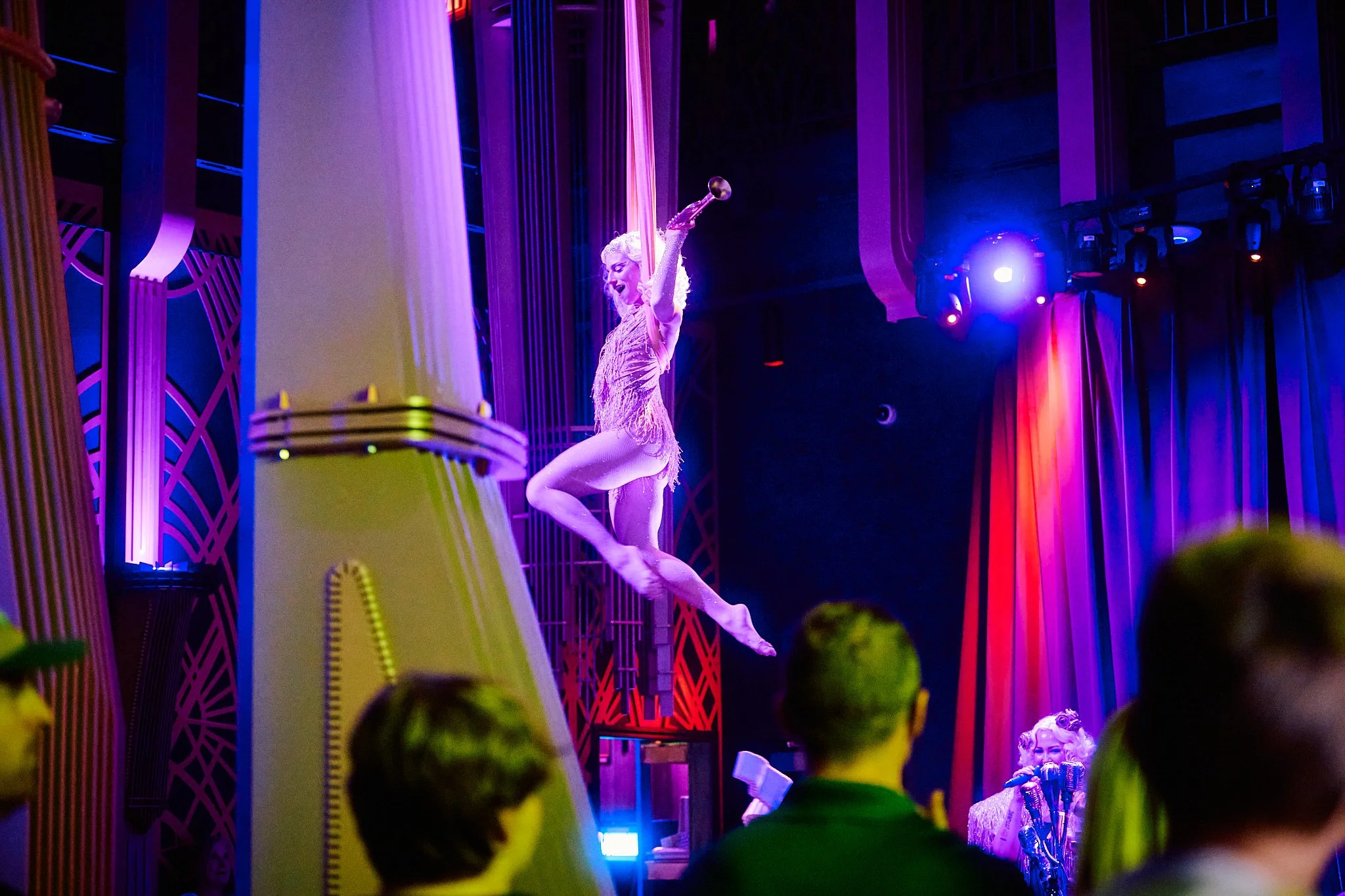 A performer in a glittery costume is on stage, jumping with one leg bent and holding a ball in one hand. The stage is lit with colorful purple, blue, and red lights, and there is an audience watching in the foreground.