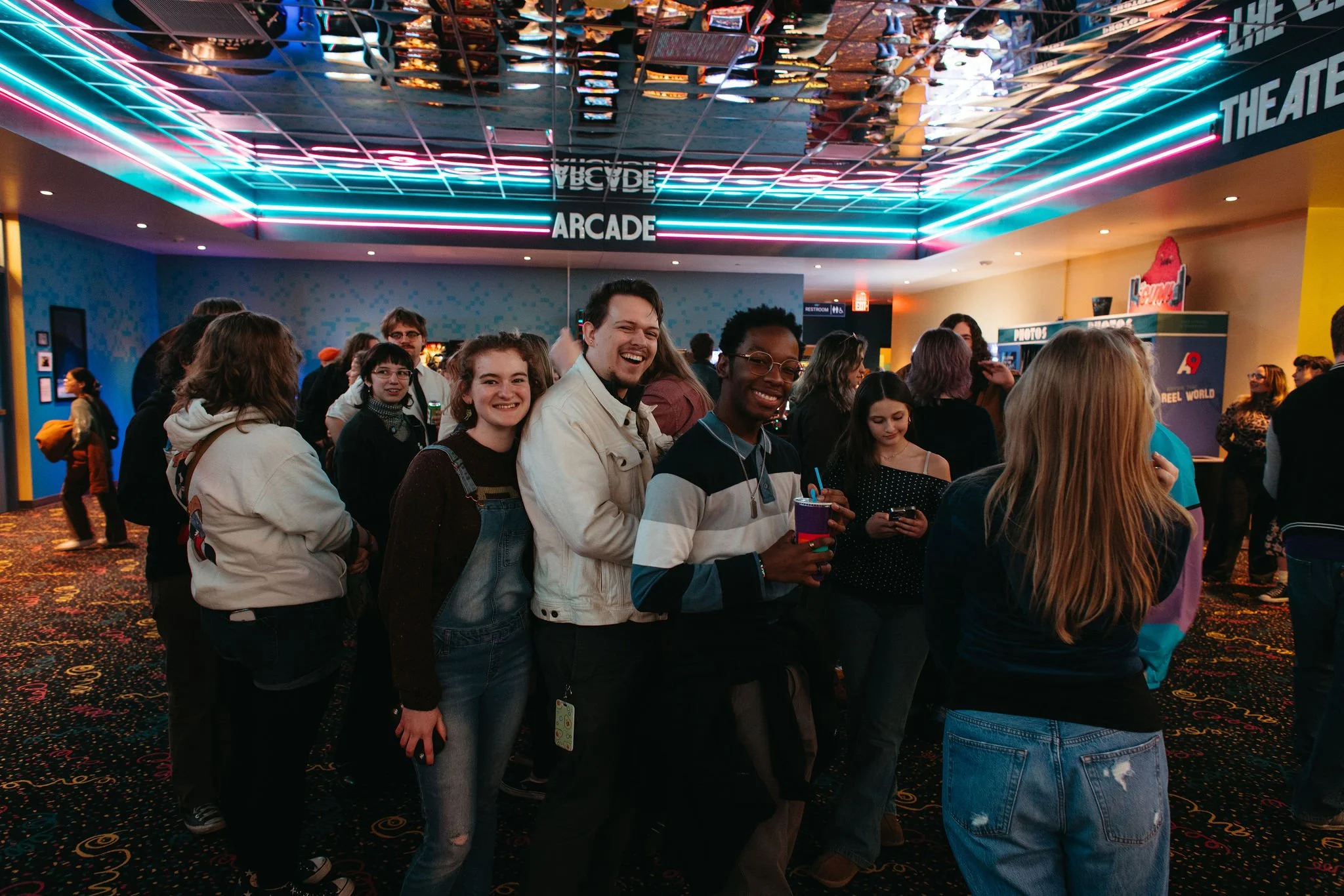 Group of people standing inside a movie theater lobby inside Atlas9 with neon lights on the ceiling, some smiling and holding drinks, others using cell phones, with signs for arcade, theater, and photo booths in the background.