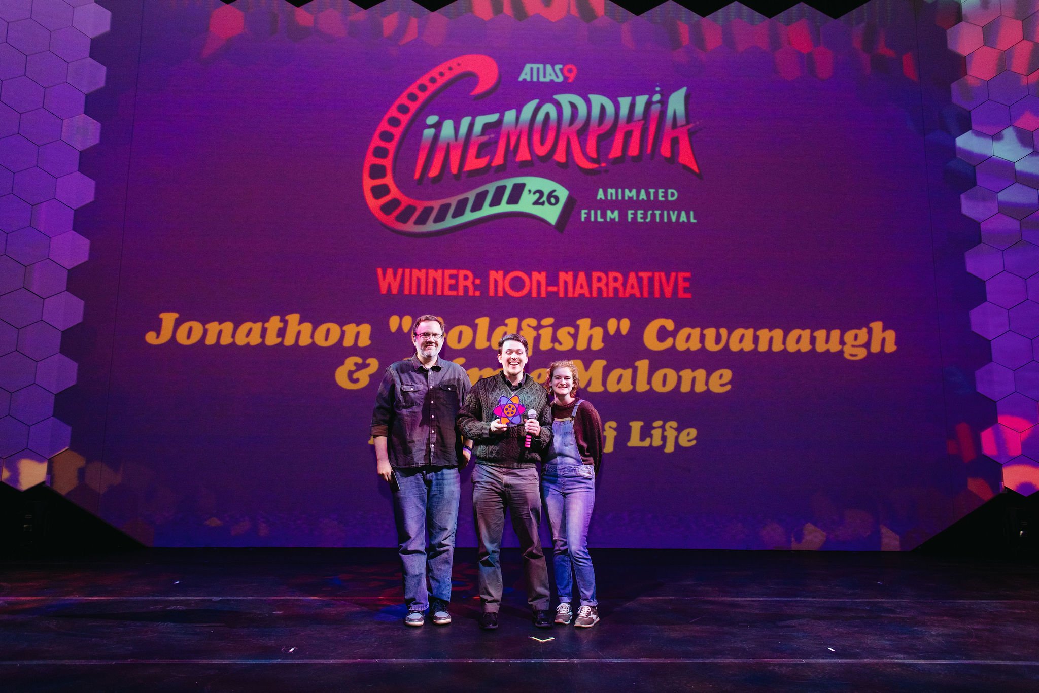 Three people standing on stage in front of a large screen displaying the winning film for the 2026 Animated Film Festival. The screen reads: 'Winner: Non-Narrative, Jonathon 'Goldfish' Cavanaugh & Emma Malone, Life'. All three are smiling, with the p