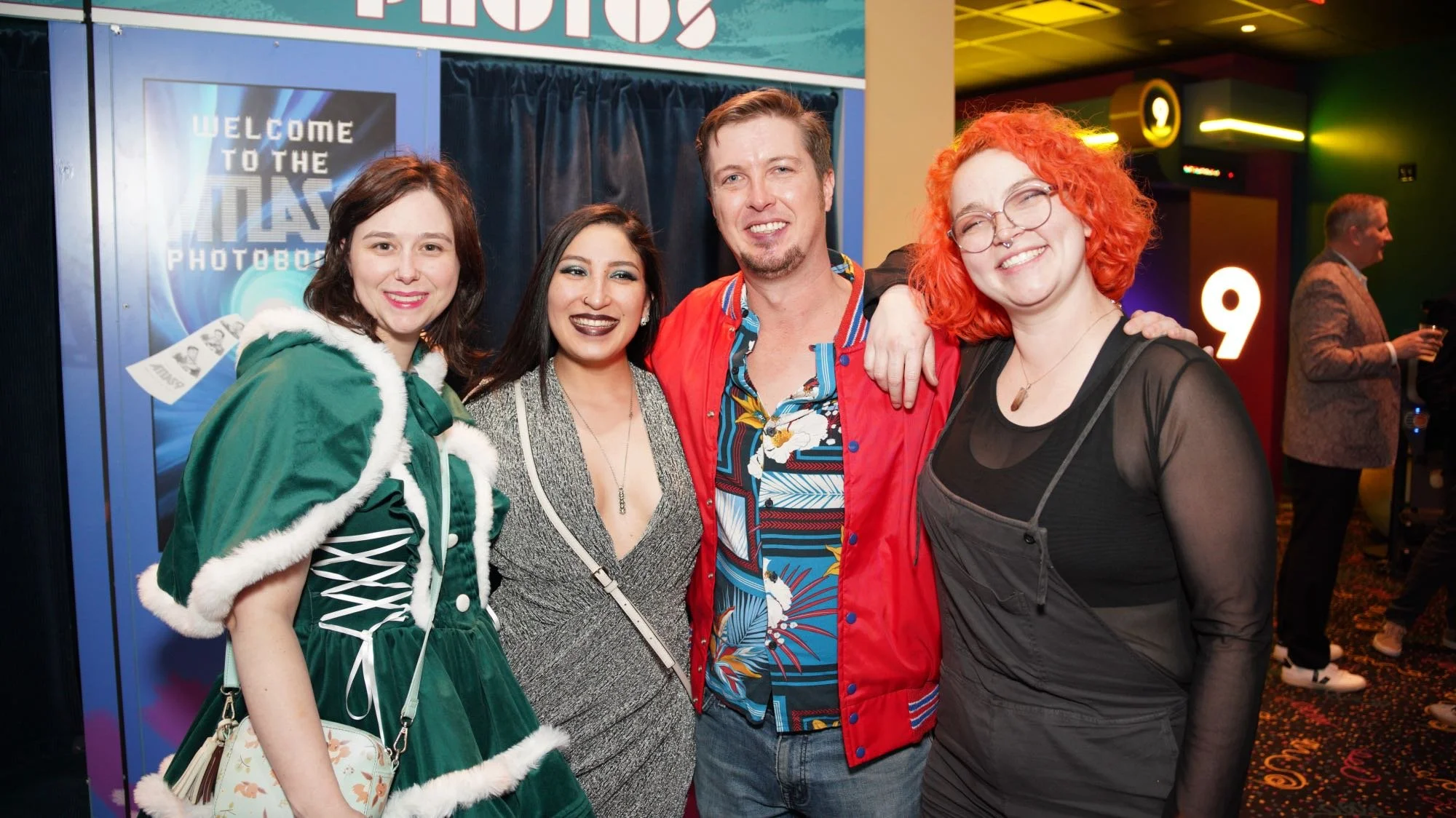 Four people smiling and posing in front of a photo booth at a party or event, with colorful lights and decorations in the background.