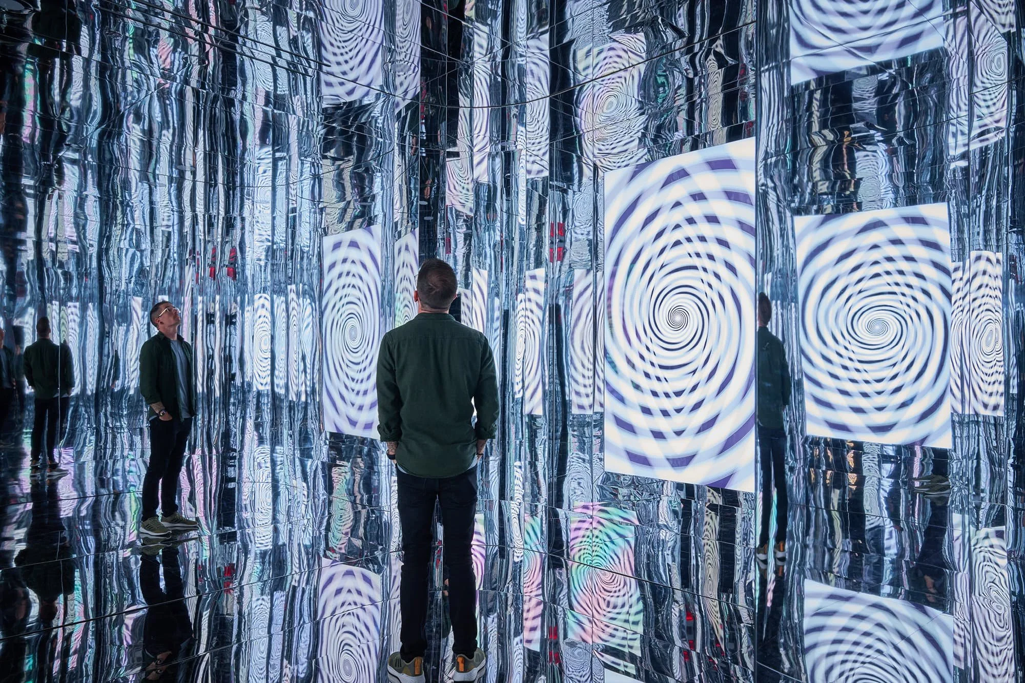 A futuristic mirrored and reflective immersive room with screens inside Atlas9. There is a man standing in the room with his reflection on all surfaces