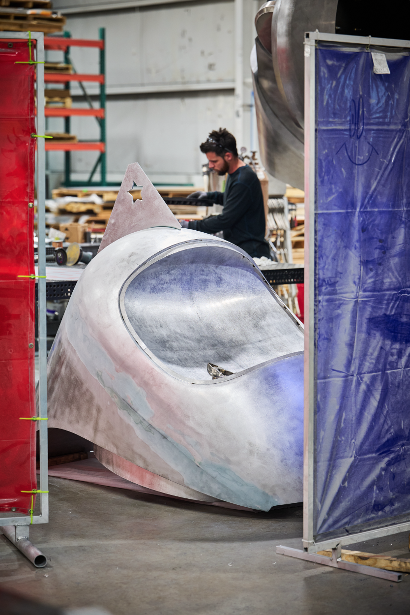 A man working on a large, metallic, unfinished sculpture resembling a spaceship or rocket for Atlas9 in a workshop at Dimensional Innovations, viewed through a red and blue protective barrier.