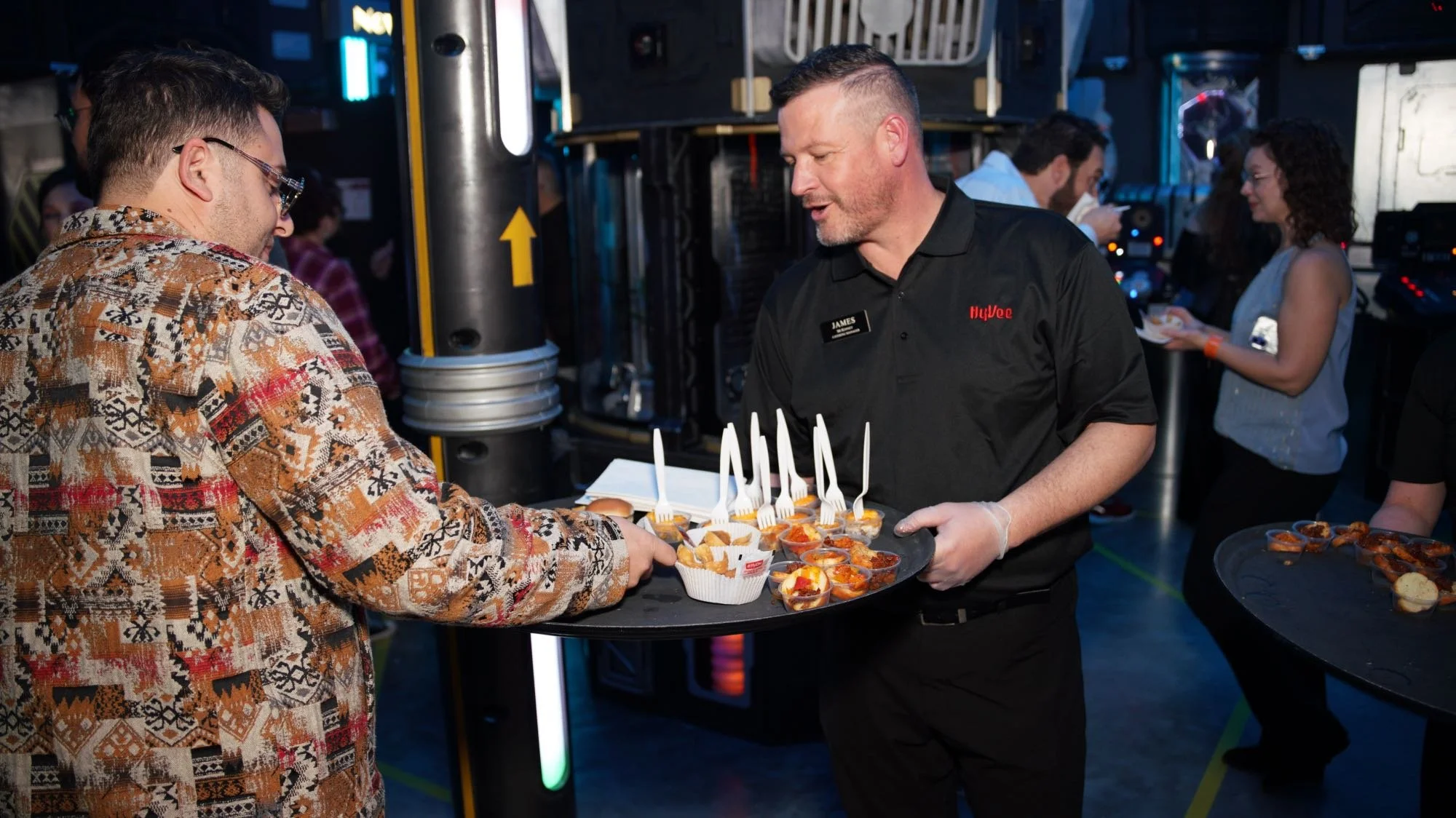 A man wearing a black shirt with a name tag that reads James and an apron serving food to a guest at a catering event. The server is holding a tray with appetizers or desserts and is handing it to a guest in a patterned shirt. There are other people 