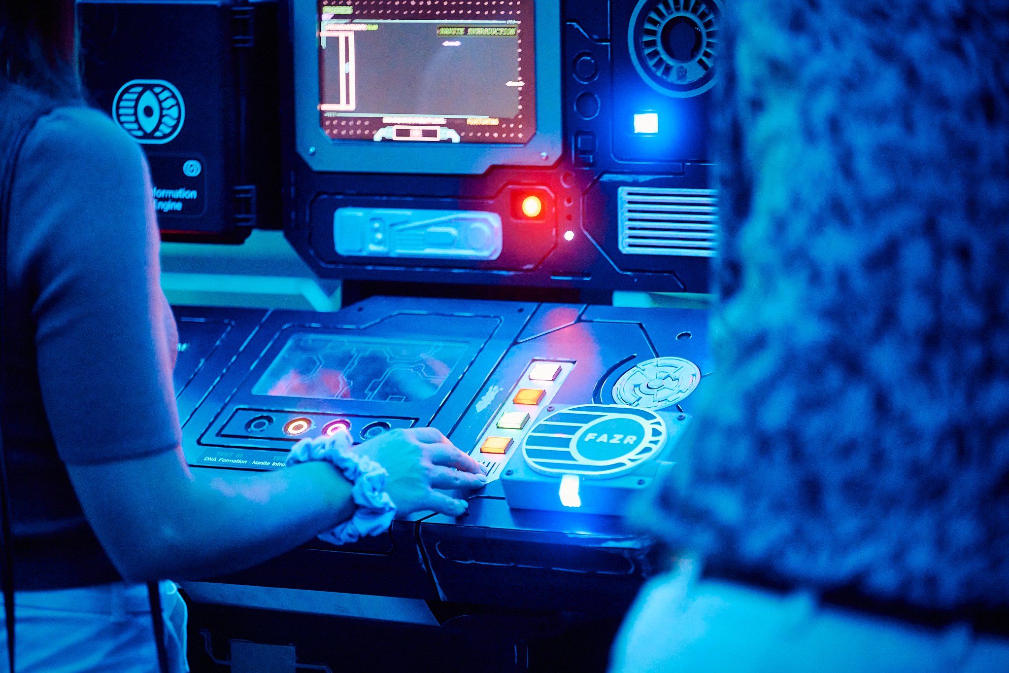 Two people are operating a futuristic control panel with colorful lights and a computer screen, in a dark room with blue lighting.