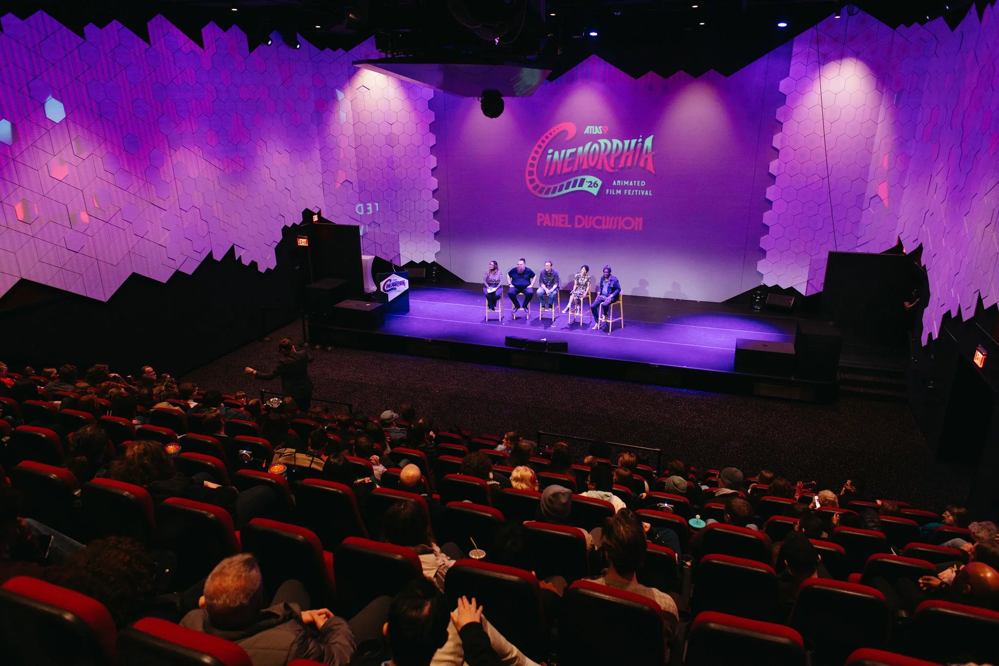A panel discussion is taking place on a stage inside Atlas9 with attendees seated in theater seats. The backdrop displays fierce purple and pink geometric designs, and the festival name includes 'Cinémorphia' with a tagline 'Animated Film Festival'.
