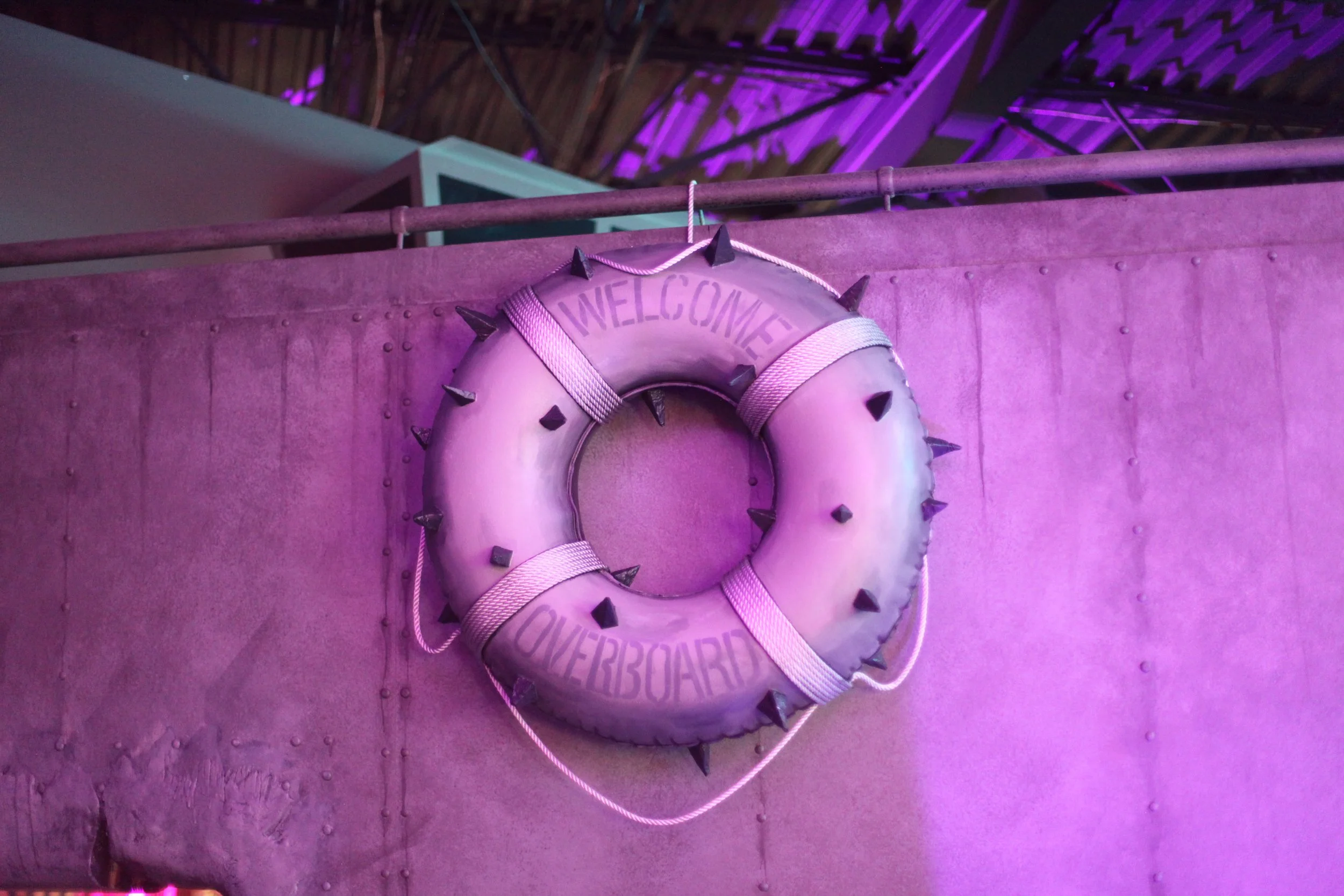 A purple life preserver with black spikes and the text 'Welcome Overboard' written on it, hanging on a rusty railing against a pink and purple background with neon lighting inside of Atlas9.