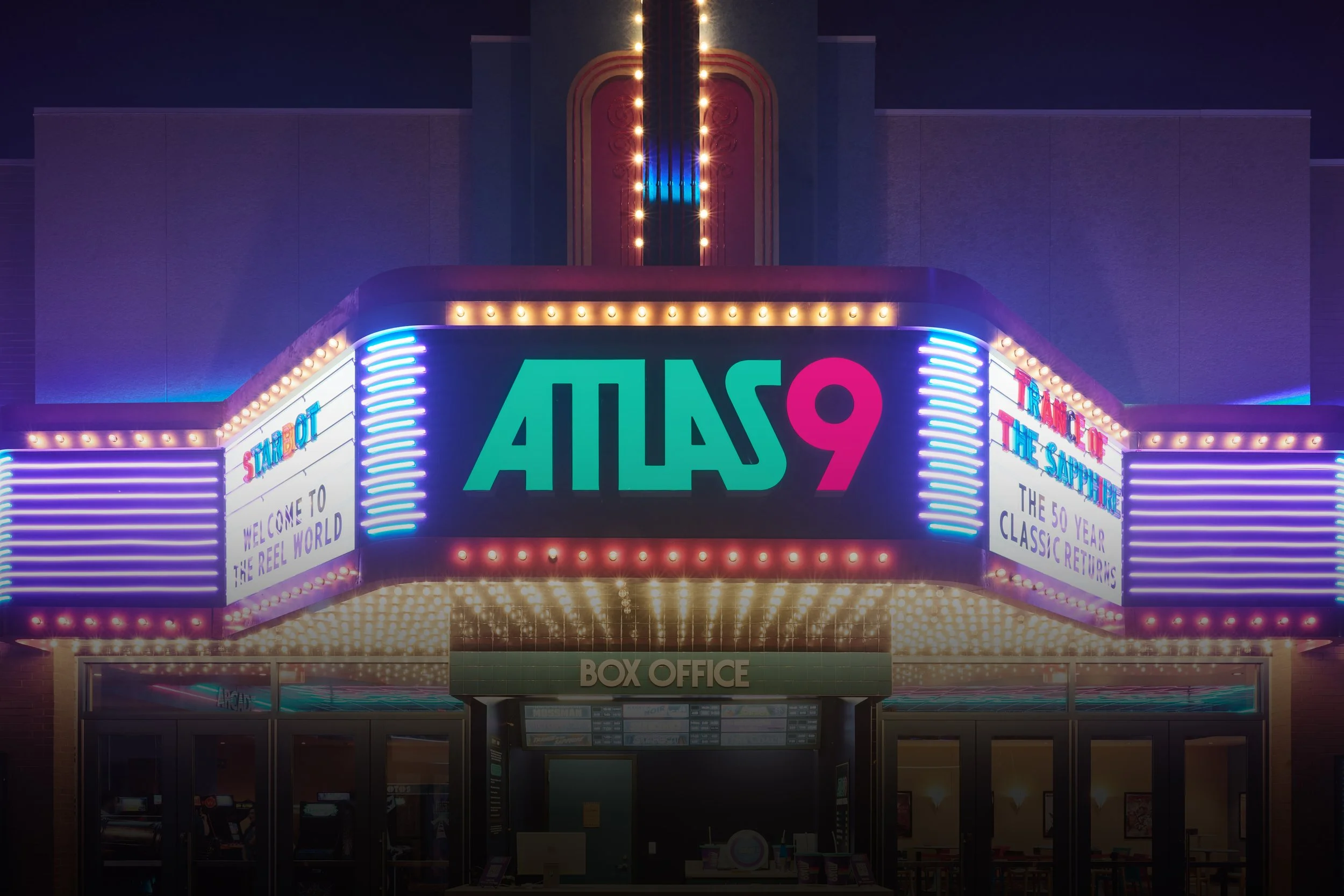 Night view of a neon-lit theater marquee at Atlas9 with bright lights and signage, featuring the words 'WELCOME TO THE REEL WORLD' and advertising classic movies.