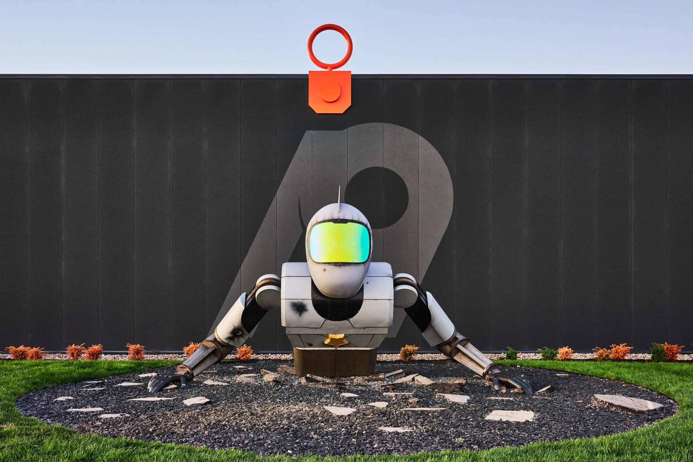 Robot in crawling position with a reflection on its visor, behind Atlas9 placed on a circular bed of rocks with grass and small bushes around, in front of a black wall with 'A9' painted on it and a red structure and hook above.