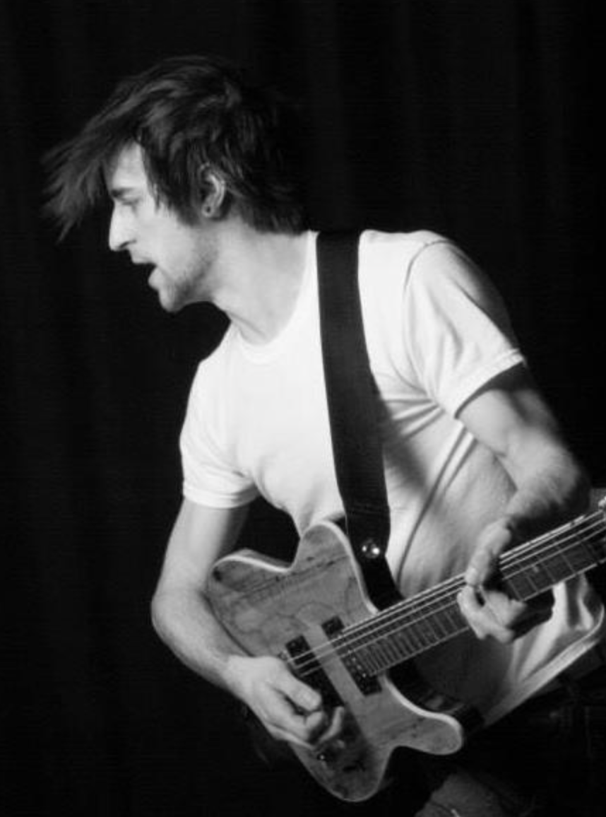 A black-and-white photo of a young man playing an electric guitar, wearing a plain t-shirt and a guitar strap, with messy hair, against a dark background.