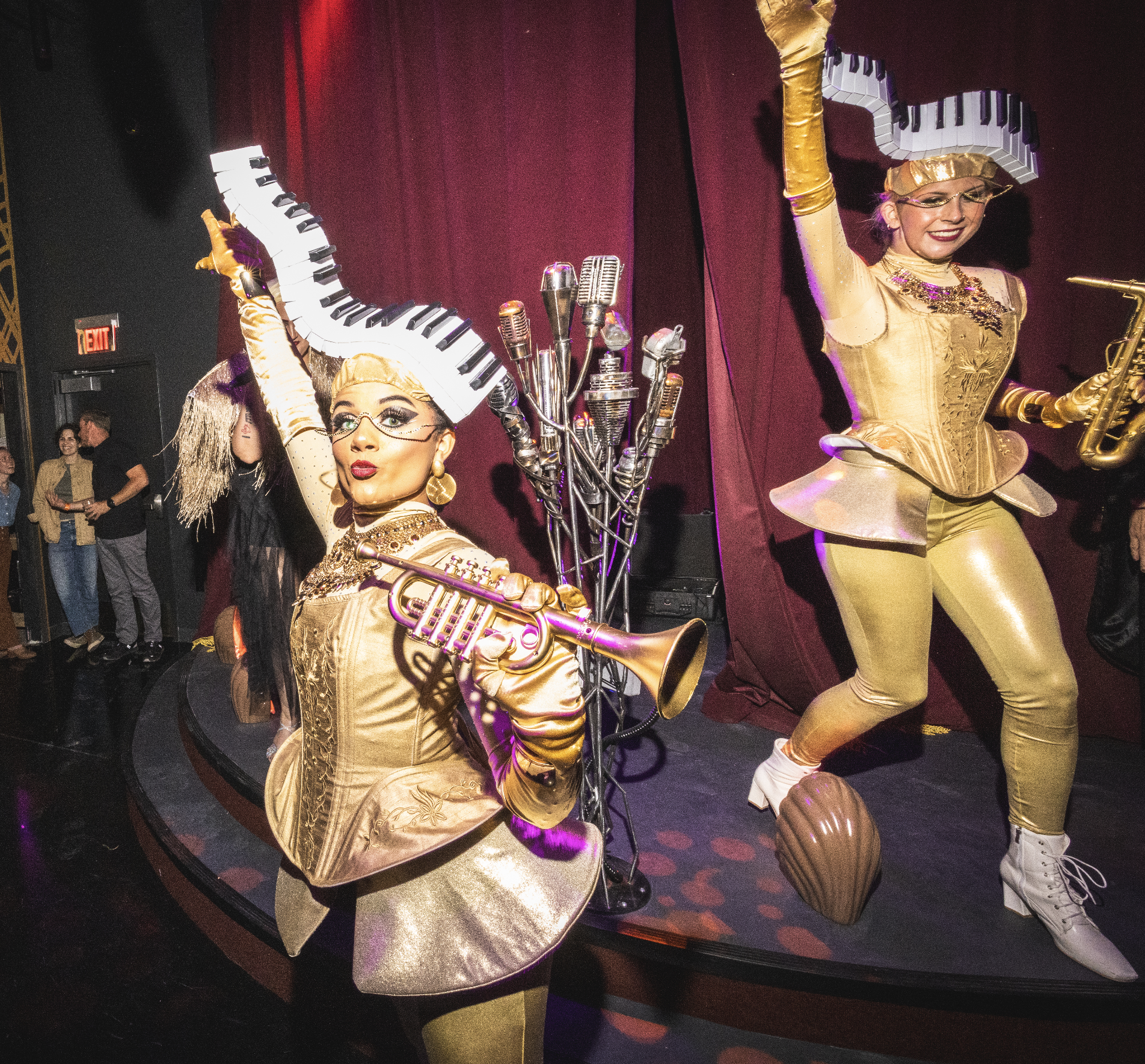Two Quixotic performers dressed in gold costumes with piano and saxophone themed accessories on stage at Atlas9, with microphones and musical instruments, in a theatrical setting.