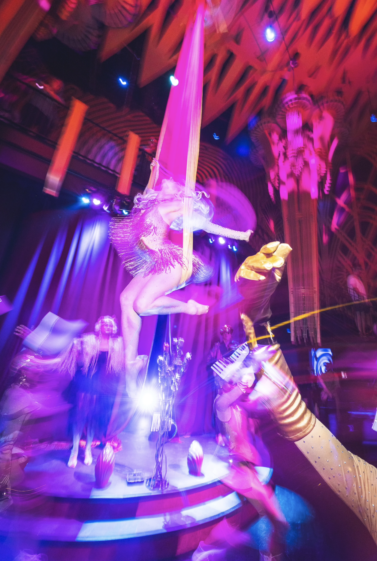 Aerial Quixotic performer in a pink and purple costume with fringe, hanging upside down from a pink silk at Atlas9 with colorful lights and a patterned ceiling.