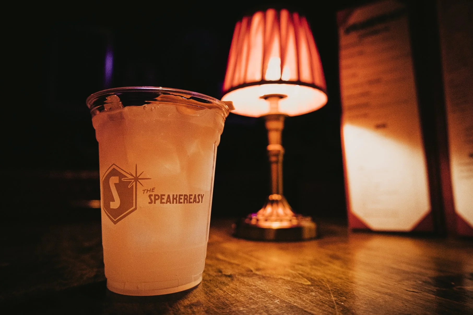 A clear plastic cup with ice and a cocktail beverage, labeled 'The SpeakerEasy,' is placed on a wooden surface. It is from Atlas9's hidden speakeasy. Behind it, there is a warm-colored table lamp with a pink lampshade and a menu or sign.