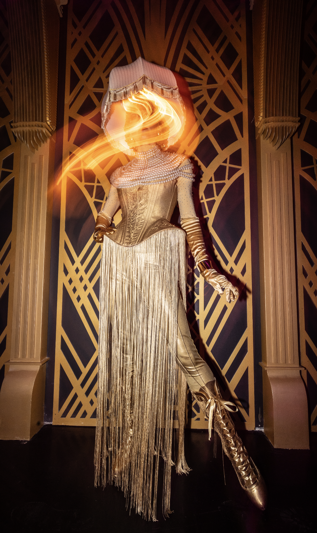 A Quixotic performer dressed in an elaborate gold costume with a fringe skirt, pearl necklace, gloves, high boots, and a large headpiece with a fringed shade, standing against a decorative gold-patterned background inside the jazz club at Atlas9.