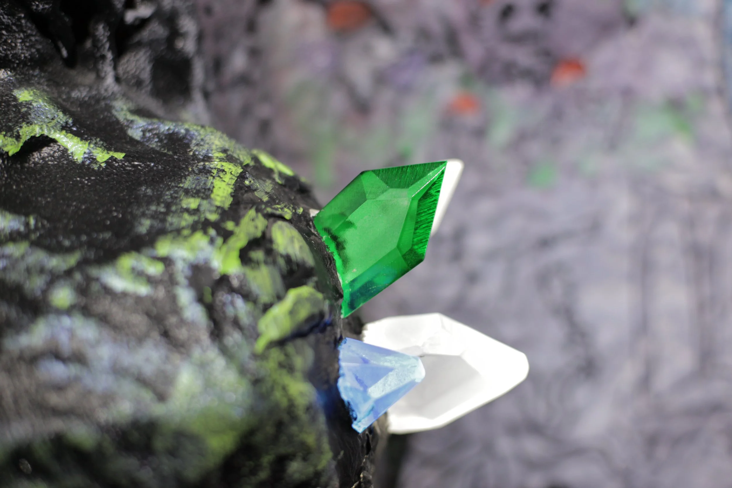 Close-up of a climbing wall with three colorful holds: green, blue, and white, attached to a textured, dark-colored rock surface inside Atlas9.