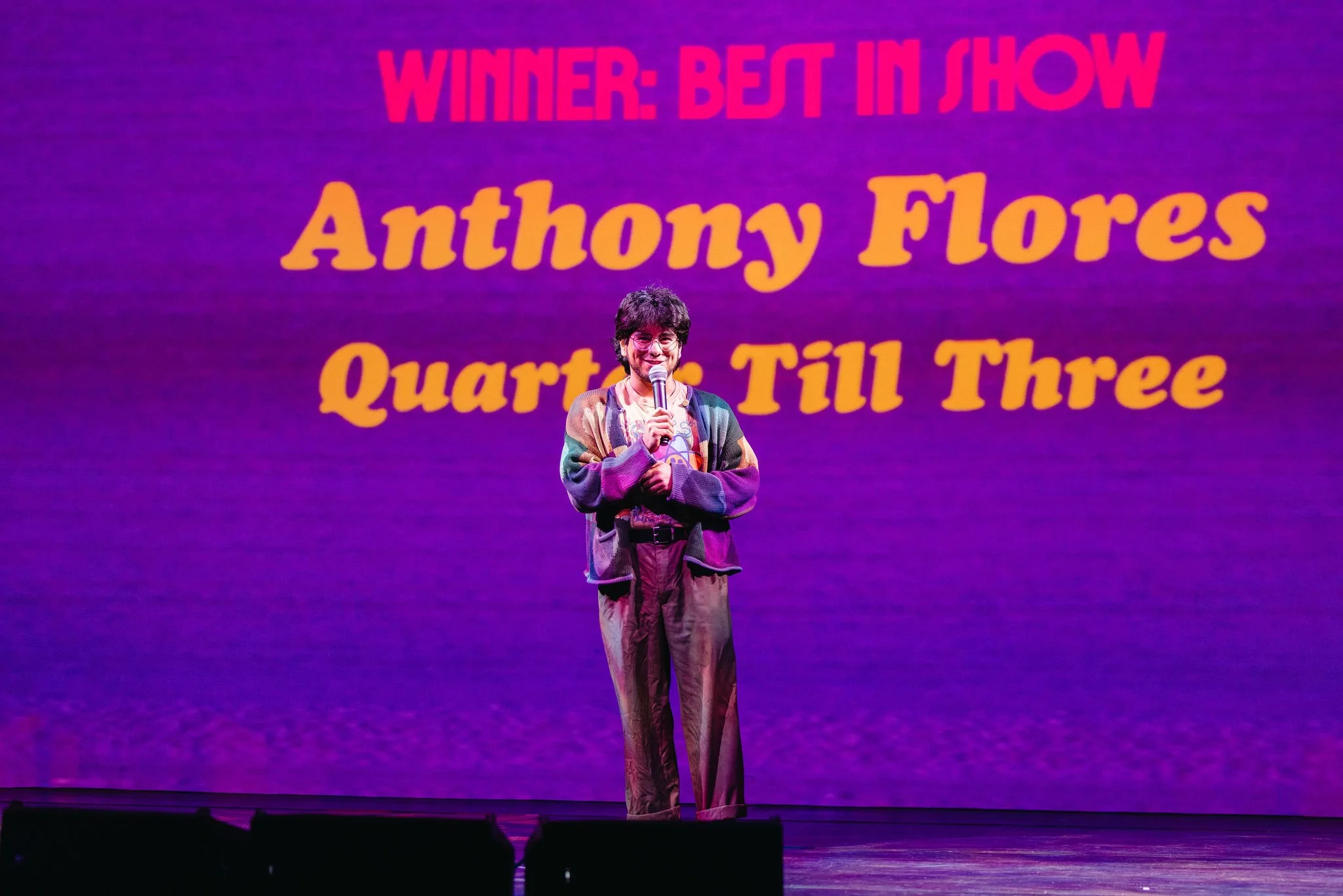 A person standing on stage holding a microphone inside Atlas9, with a large screen behind them displaying text that reads "WINNER: BEST IN SHOW Anthony Flores Quartet Till Three" in bright colors.