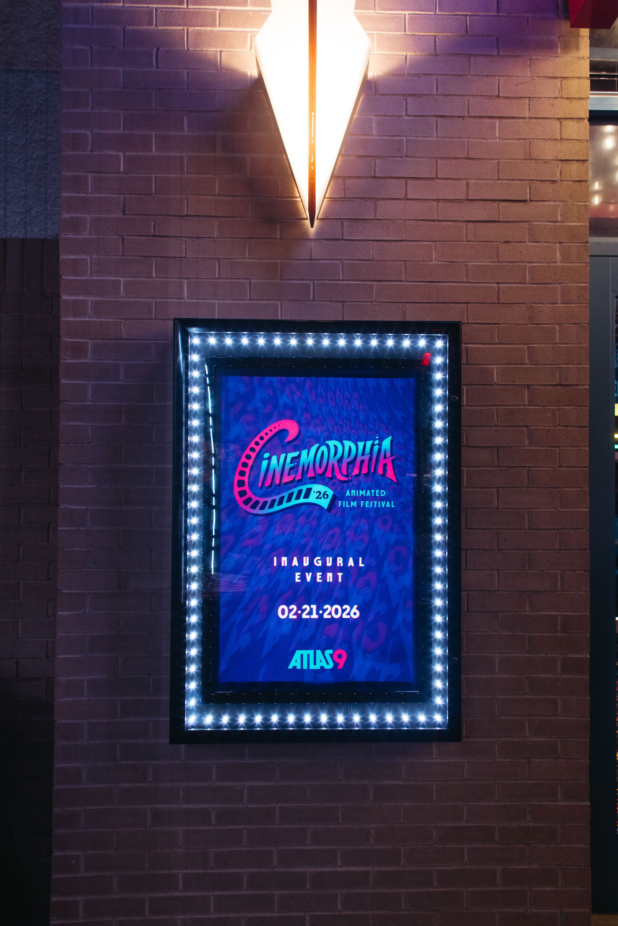 Digital poster for Cinemorphia 26 animated film festival, showing inaugural event date February 21, 2026, with a neon-style design and illuminated border, mounted on a brick wall below a geometric wall sconce inside Atlas9.