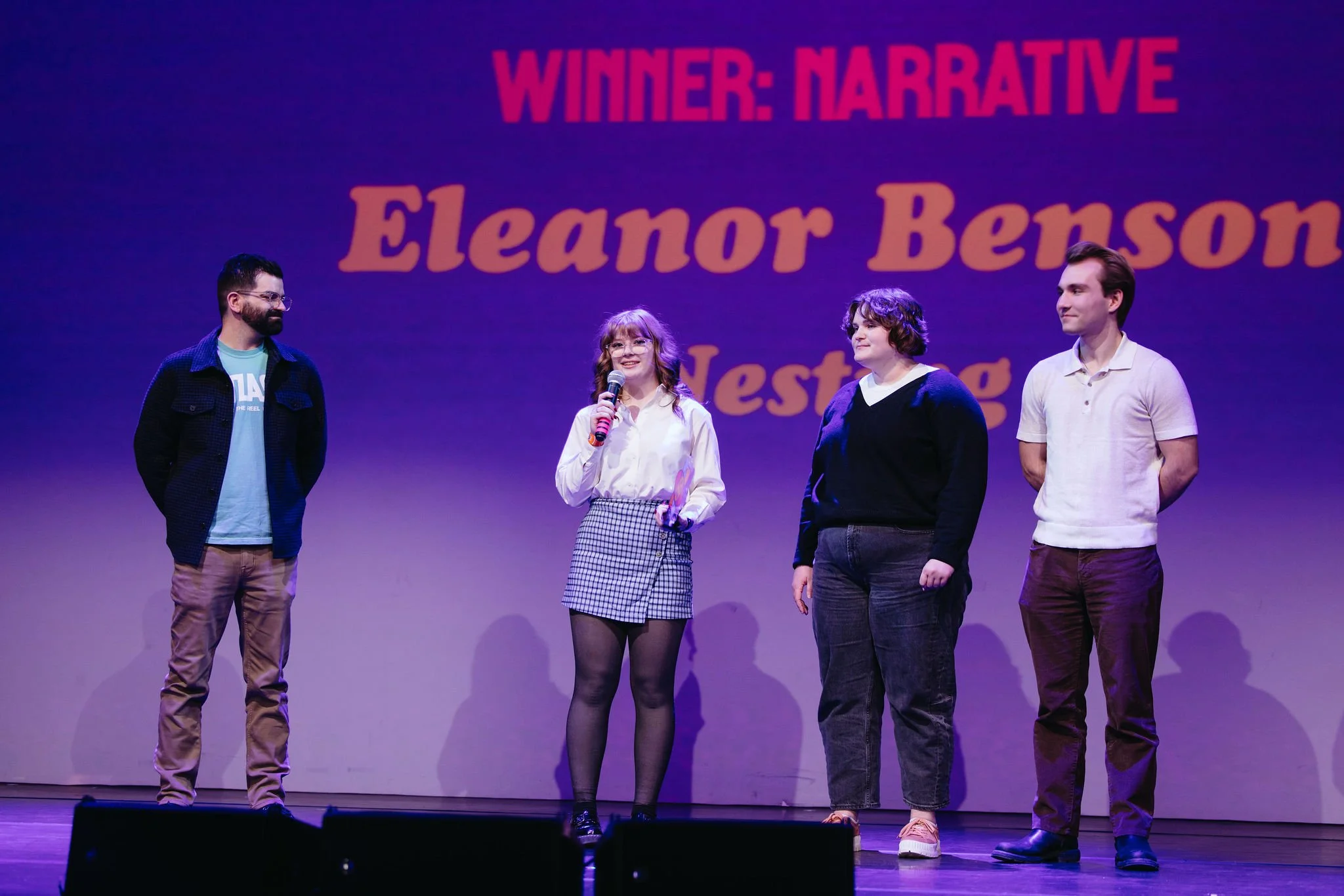 Four people stand on stage inside Atlas9 during an award ceremony, with a woman in the center speaking into a microphone. Behind them is a large screen showing text that reads 'WINNER: NARRATIVE Eleanor Benson.' The stage is lit with purple lighting.