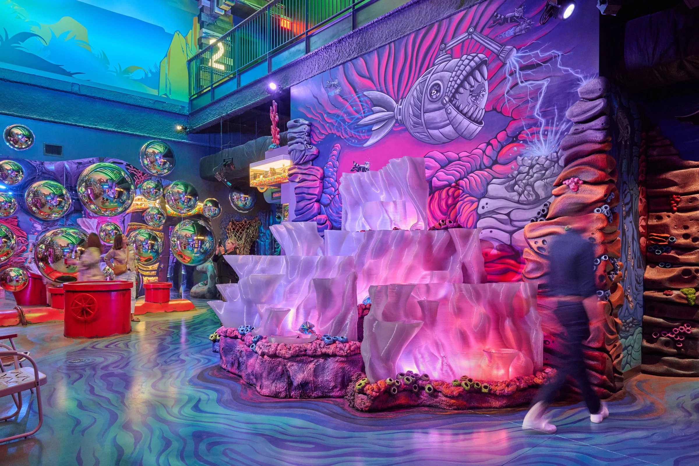Interior  with vibrant neon lighting, colorful mural art featuring a fish with lightning, reflective balls on the wall, and a cascade-shaped structure illuminated with pink and purple lights. People are seen in the background playing games and walkin