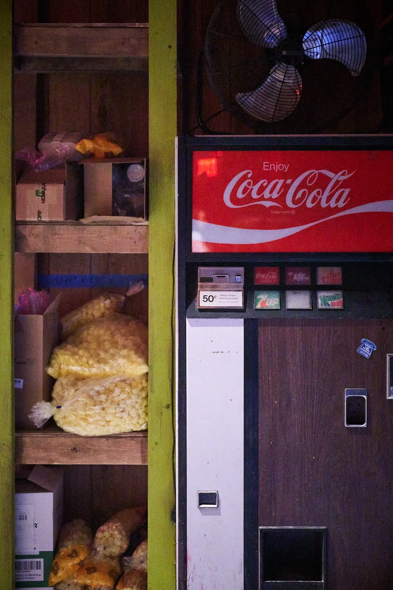 A vending machine with a Coca-Cola sign and a display for coins and bills, next to a wooden shelf with bags of popcorn and other snacks.