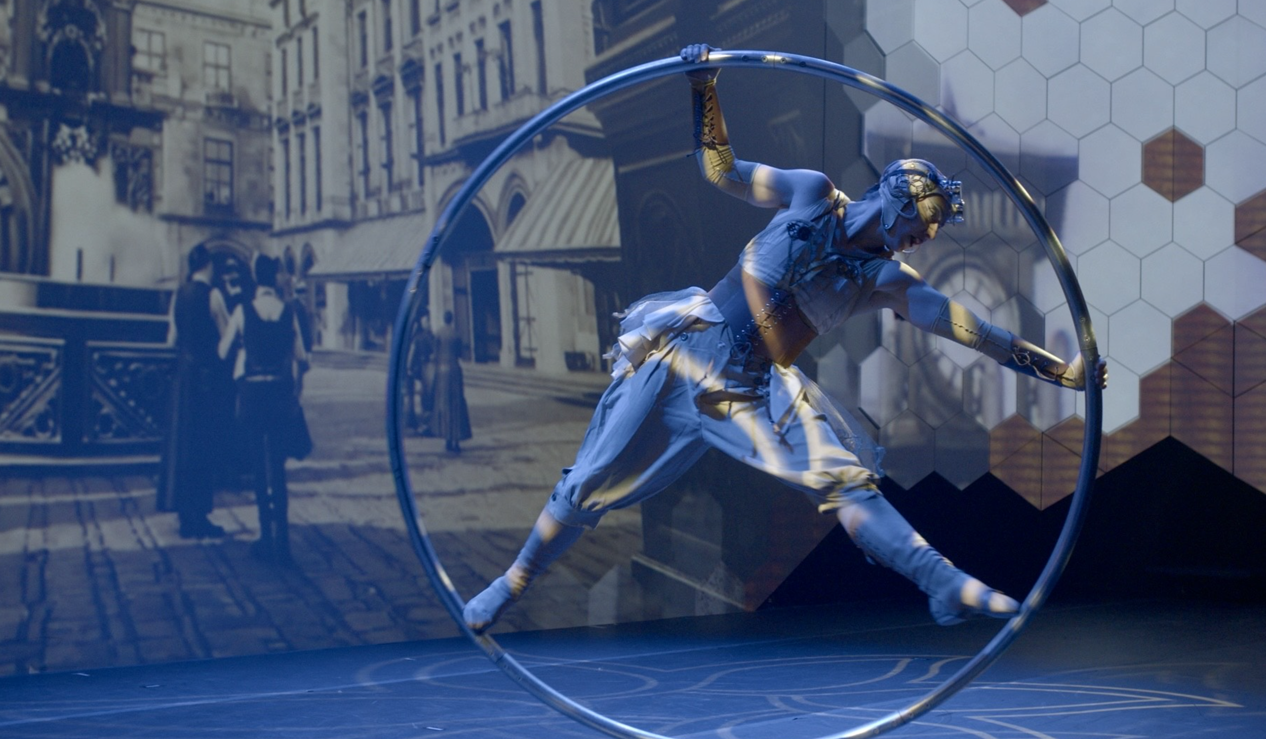 A Quixotic performer dressed in futuristic attire twirls inside a large metal ring on a stage at Atlas9. The background features a black-and-white urban scene projected on the wall.